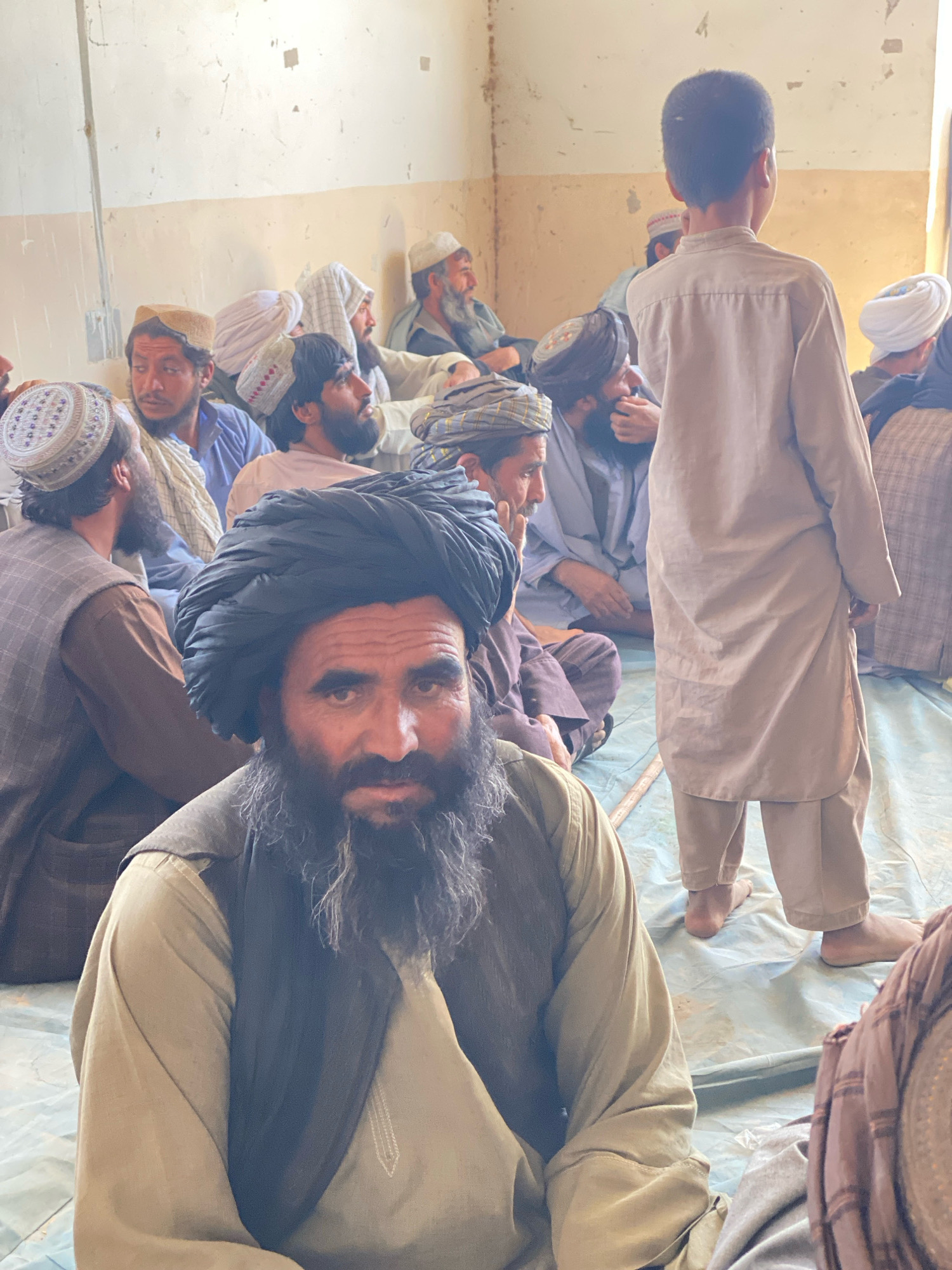In an indoor setting, likely a room in Pasab, Afghanistan, a group of men are gathered, seated on the floor. The walls of the room are pale and weathered, with visible signs of age and perhaps some discoloration. The floor is covered by a light-colored tarp or sheet.
In the foreground, a man with a prominent black turban and a long, graying beard is looking directly at the camera. He is wearing a light-colored shirt and a dark vest or shawl. To his left, another man is also seated, wearing a light brown vest over a light blue shirt and a patterned cap. His head is turned to the side.
In the mid-ground, a young boy stands with his back to the camera, wearing a light-colored tunic and trousers. Behind him and to his right, several other men are visible, some wearing turbans and traditional clothing. One man in the mid-ground, wearing a white turban, appears to be speaking or looking towards someone off-camera.
The overall atmosphere suggests a gathering or meeting, possibly a community or religious event. The lighting is natural and somewhat diffused, indicating it is likely daytime. The scene is candid, with no clear signs of posed activity.