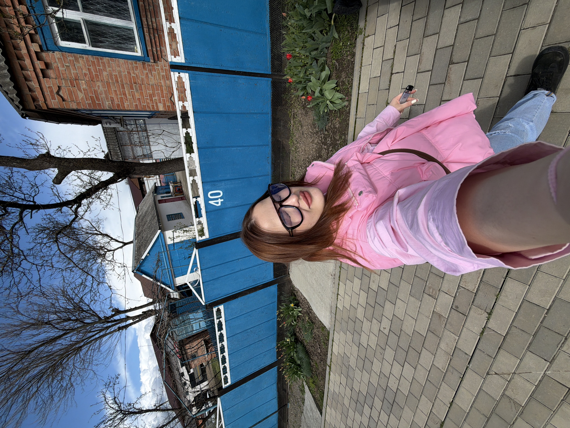A young woman wearing glasses, a pink jacket, and light-wash jeans poses for a selfie on a paved walkway next to a blue fence with the number "40" on it. She is holding a small electronic device in her right hand. A row of flowers and foliage borders the walkway and fence. Behind the fence is a view of residential houses, including a brick building and a blue-painted structure, all beneath a clear sky with the bare branches of trees visible.