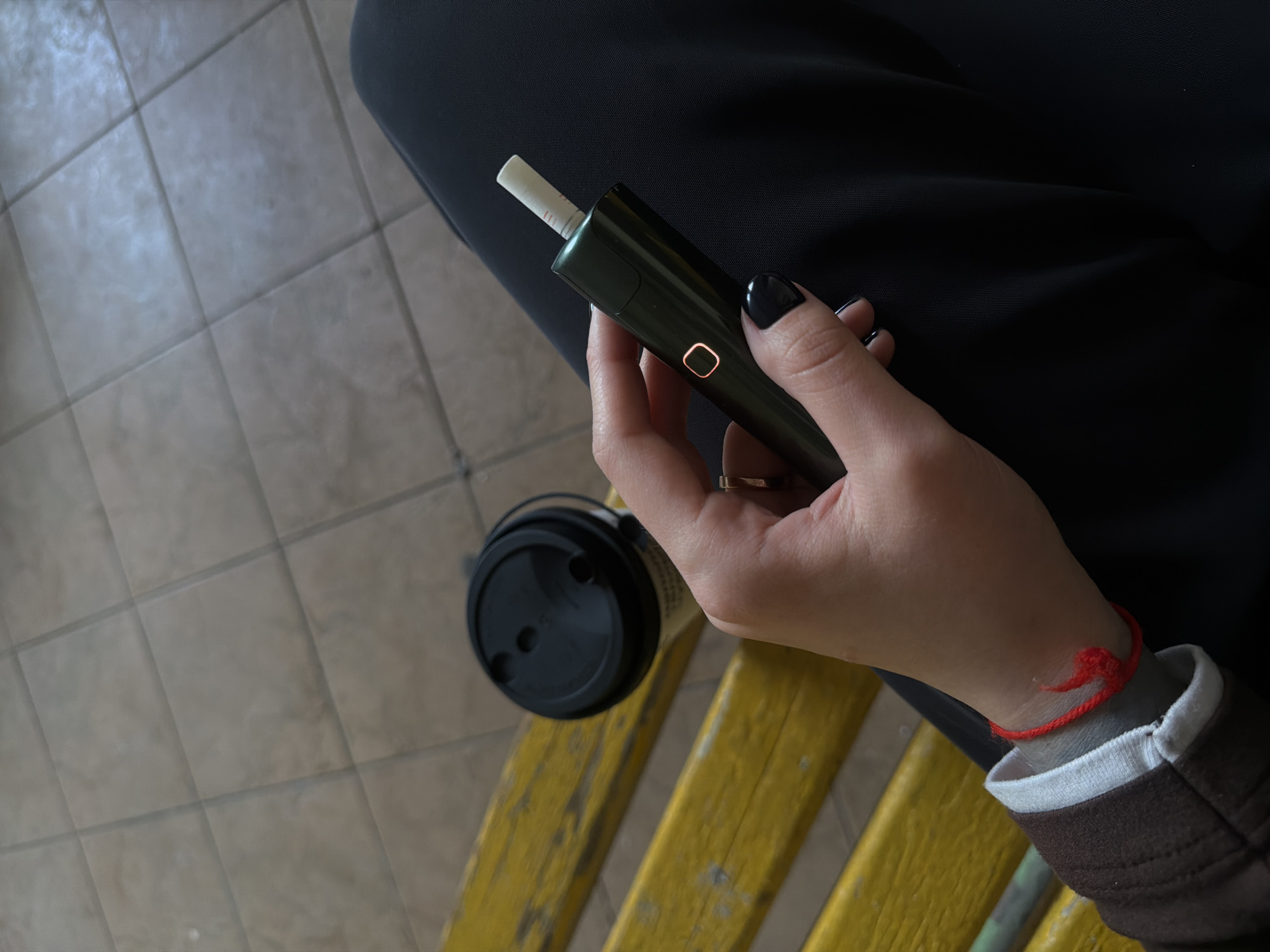 A close-up shot focuses on a person's hand holding a dark green electronic smoking device. The device has a glowing orange square indicator light. A white cigarette-like stick protrudes from the top of the device. The hand has black nail polish and a ring on the index finger. The person is wearing a brown jacket with white trim and a red string bracelet on their wrist. To the left of the hand, a black-lidded paper cup is visible, resting on a bright yellow surface. The background shows beige tiles on the floor and a dark fabric, likely clothing. The scene appears to be indoors or in a sheltered outdoor area, possibly a waiting room or public space. The lighting suggests daytime.