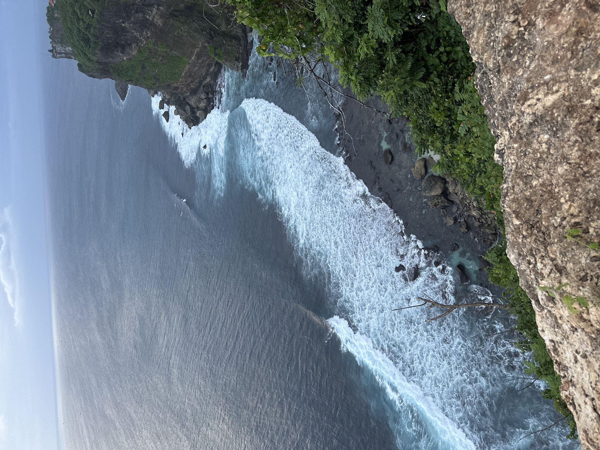 An elevated, birds-eye, sideways-oriented shot shows a section of coastline in Kuta Selatan, Indonesia. The ocean covers the majority of the view, appearing as a dark, reflective body of water. Whitewater waves are seen crashing near the rocky shore. The cliff face is marked by vegetation and rough, textured rock formations.