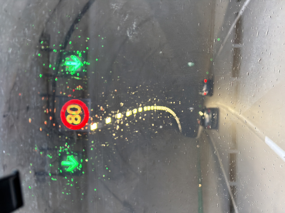 The image depicts a forensic view from inside a vehicle looking forward through a wet windshield, specifically within a tunnel located in the city of Terjola, Georgia. The windshield is obscured by numerous water droplets, causing light sources to appear as blurred bokeh.
In the upper-center, a circular red speed limit sign displays "80" in yellow characters. To the left and right of this sign, two green downward-pointing arrow indicators are visible. The tunnel's interior is illuminated by overhead lights, visible as a curving, segmented line of yellow bokeh on the upper left, and reflecting on the wet road surface below.
Two dark-colored passenger vehicles are observed ahead in the tunnel, traveling in the same direction as the observer. Both vehicles have illuminated red taillights. The roadway is marked with a solid white line on the left and a dashed white line on the right, indicating traffic lanes. The environment is a concrete-lined tunnel, with visible road infrastructure. No individuals are directly discernible.