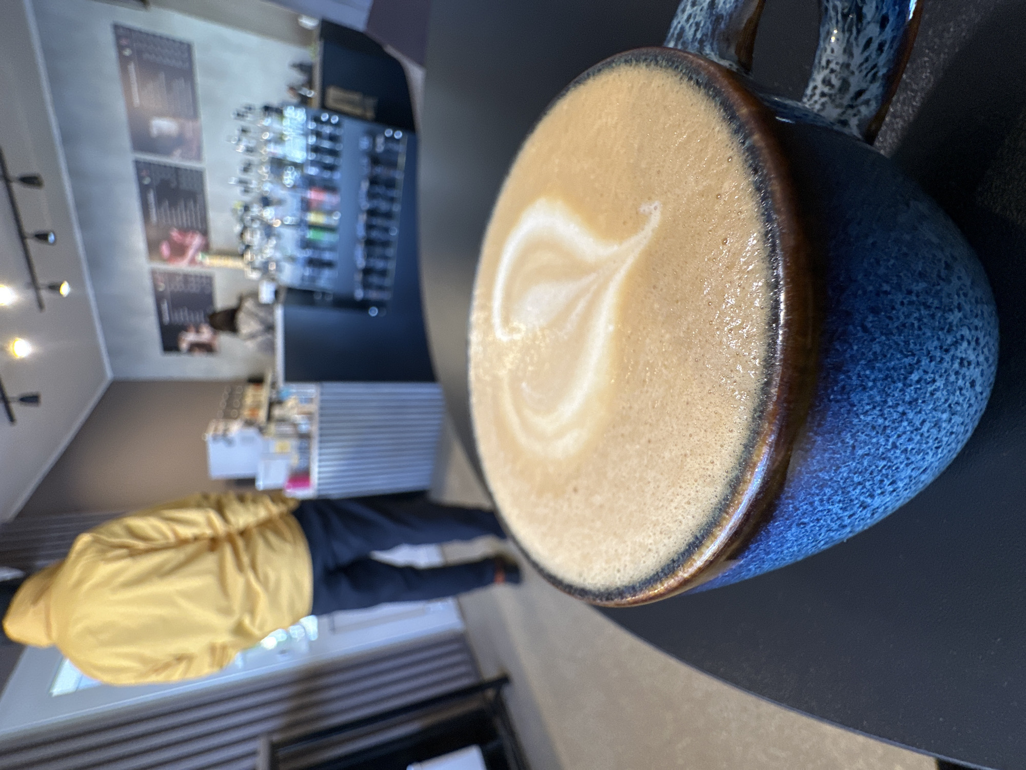 A latte art coffee is in the foreground in a blue speckled ceramic mug with a brown lip. The mug sits on a dark gray surface. A person in a yellow jacket and dark pants stands by a counter in the background. Behind the counter is a display of goods. The image was taken indoors. The general location is inferred to be Chaikovsky, Russia.