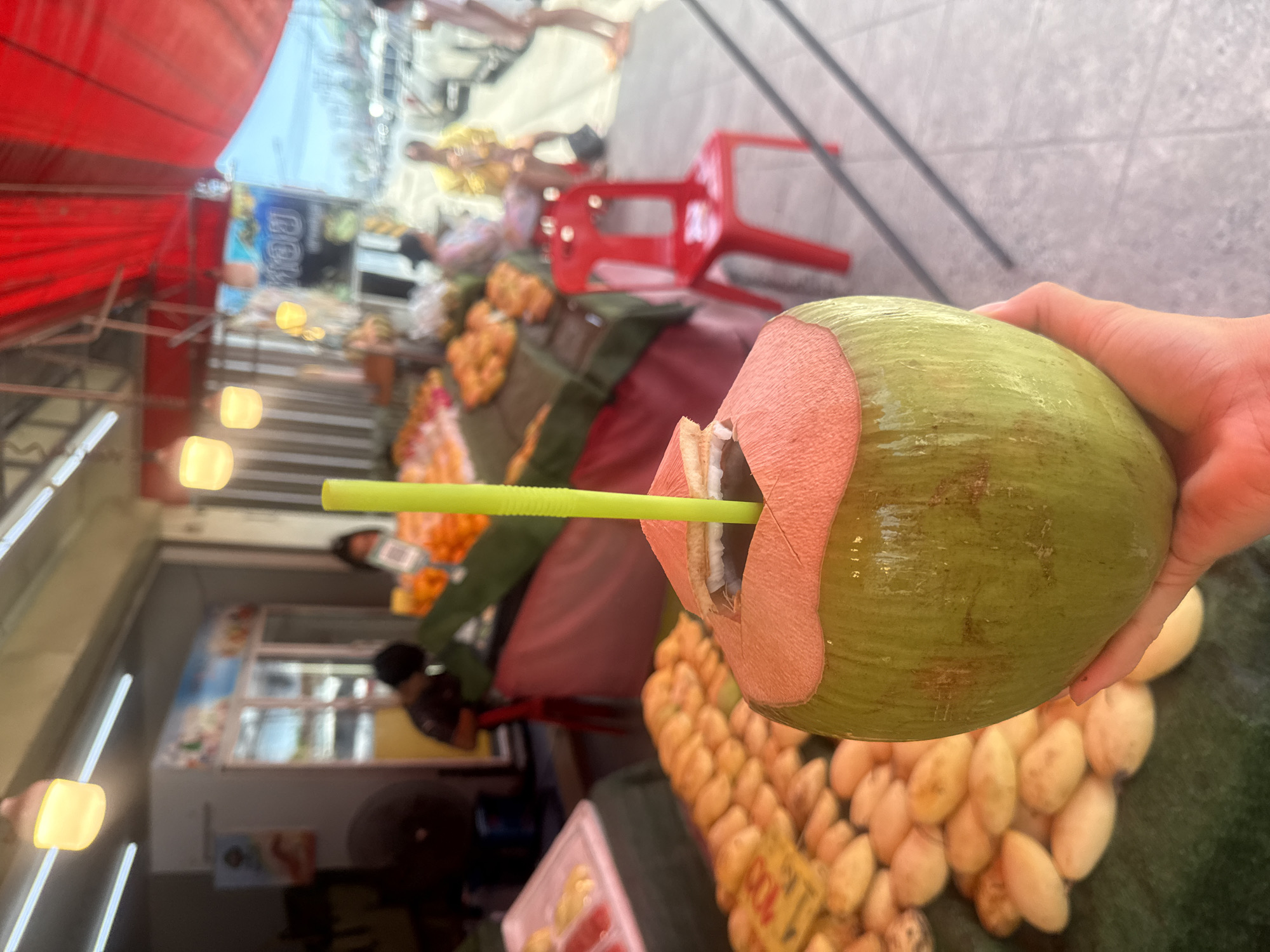 A person's hand holds a freshly cut green coconut with a bright yellow straw inserted into it. The coconut is positioned in the foreground, with its top opening displaying white flesh. In the background, a market scene unfolds. Stalls are laden with various fruits, including piles of mangoes and oranges. Several people are visible, some tending to the stalls and others browsing or sitting. Red awnings are stretched over the market area, providing shade. To the right, a red plastic chair sits on a paved walkway. The environment suggests a bustling outdoor market in Ko Samui District, Thailand, likely during daytime given the bright lighting.