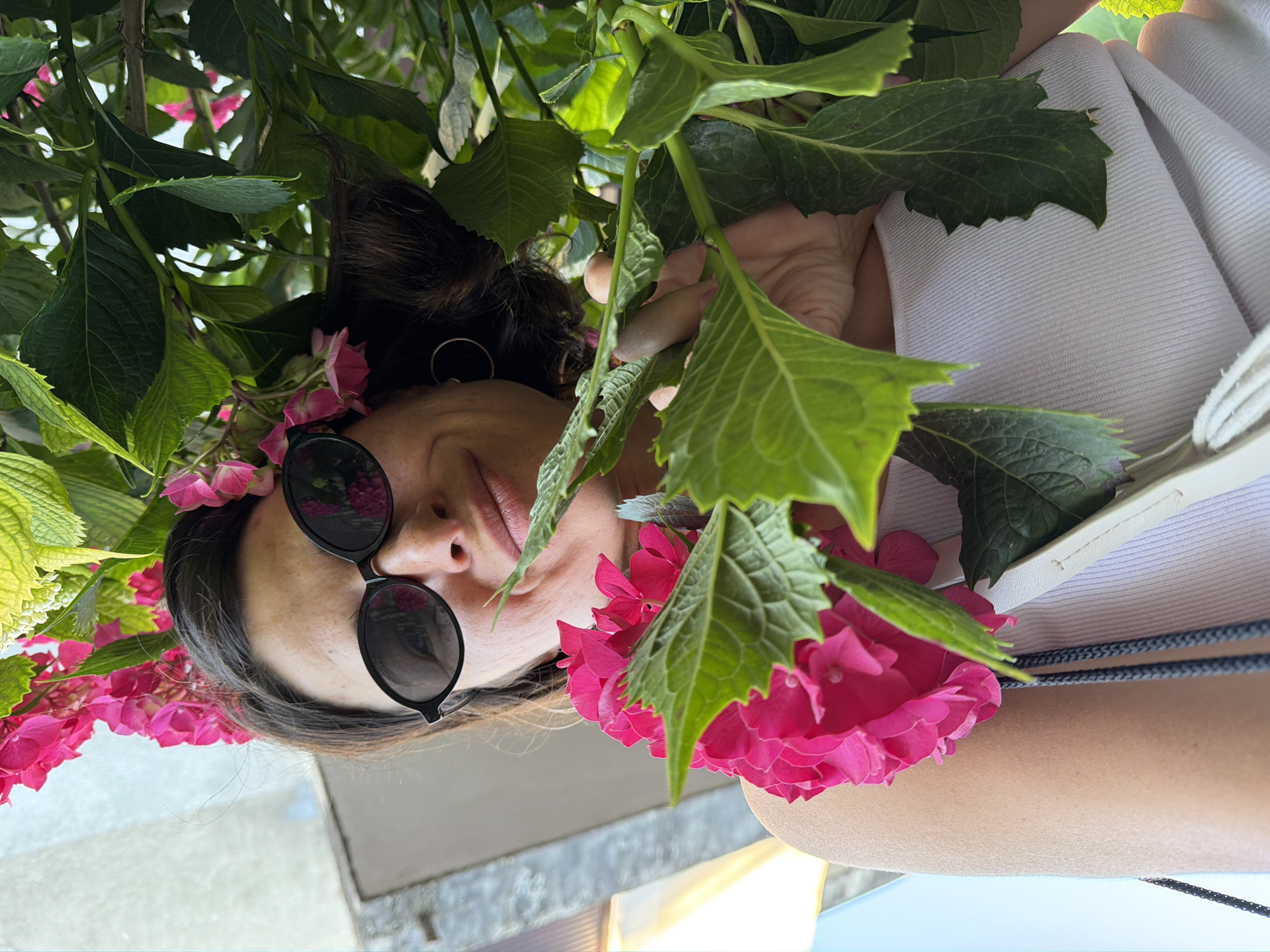A woman with sunglasses is partially visible amidst green leaves and pink hydrangea flowers. She wears a white top and earrings. The location is Budva, Montenegro. The background includes a stone surface and light blue sky.