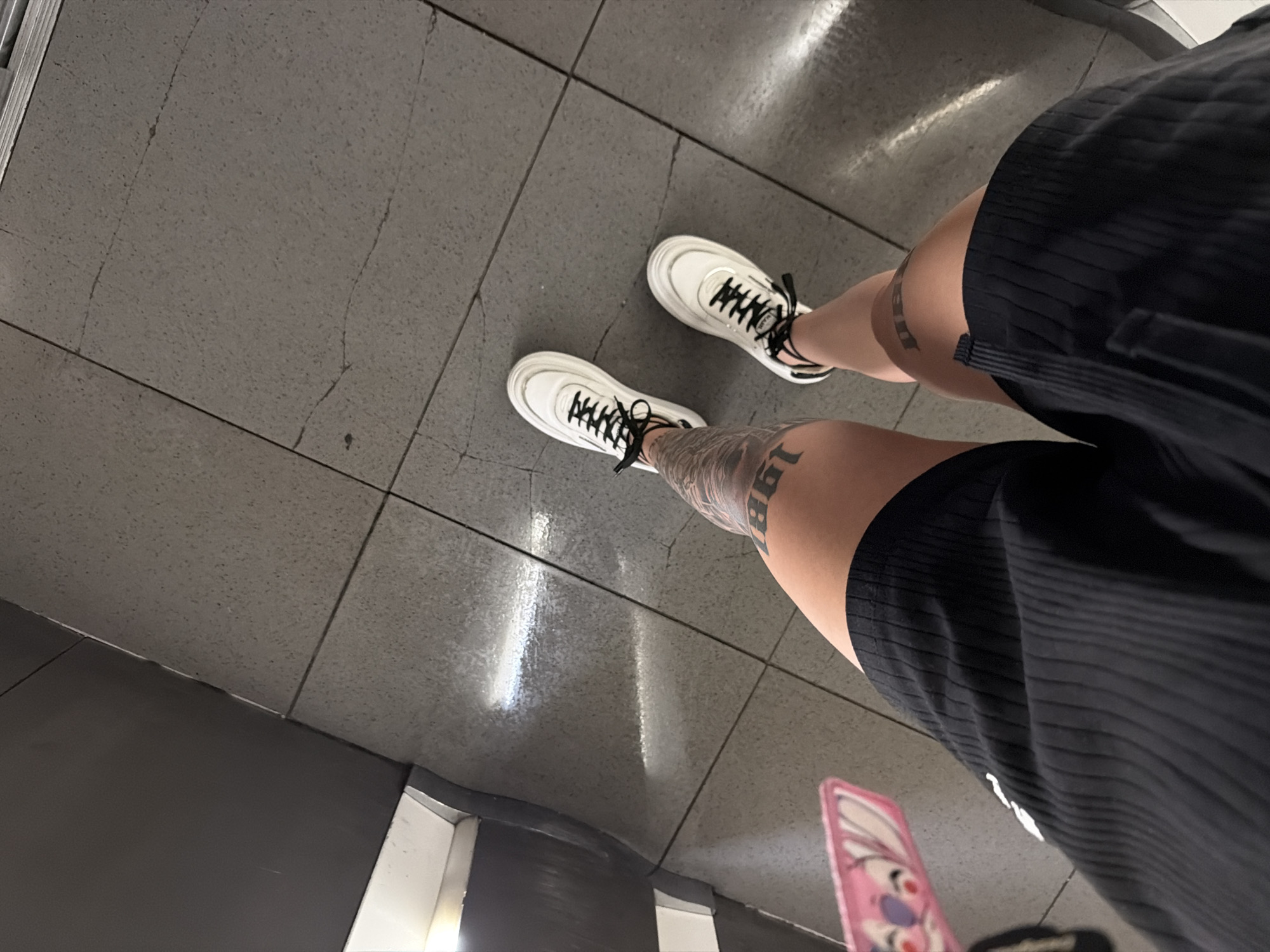 The image is a first-person, downward-angle perspective, showing the lower body of an individual standing on a tiled floor. Two legs are visible, wearing black ribbed shorts. Both legs have prominent dark tattoos; the left leg (viewer's left) displays large numbers, partially obscured, appearing to be "198", while the right leg (viewer's right) shows dark lettering on the thigh, partially obscured.
The individual is wearing white platform sneakers with thick white soles and black laces. The brand name "PRADA" is visible on the laces of the left shoe.
The floor is composed of large, light grey, speckled tiles with dark grout lines. Several thin, dark cracks are visible across the tile surfaces. To the left, a dark grey, reflective vertical surface meets the tiled floor, with a white horizontal surface visible beneath it, suggesting a baseboard or ledge. Strong, elongated reflections of artificial light sources are present on the glossy dark grey surface and the adjacent floor tiles. A small, partially visible pink object with a cartoon character (resembling Stitch) is present in the lower left corner of the frame, possibly part of an accessory. There are no discernible elements within the image to indicate a specific location in the city of Plovdiv, Bulgaria.