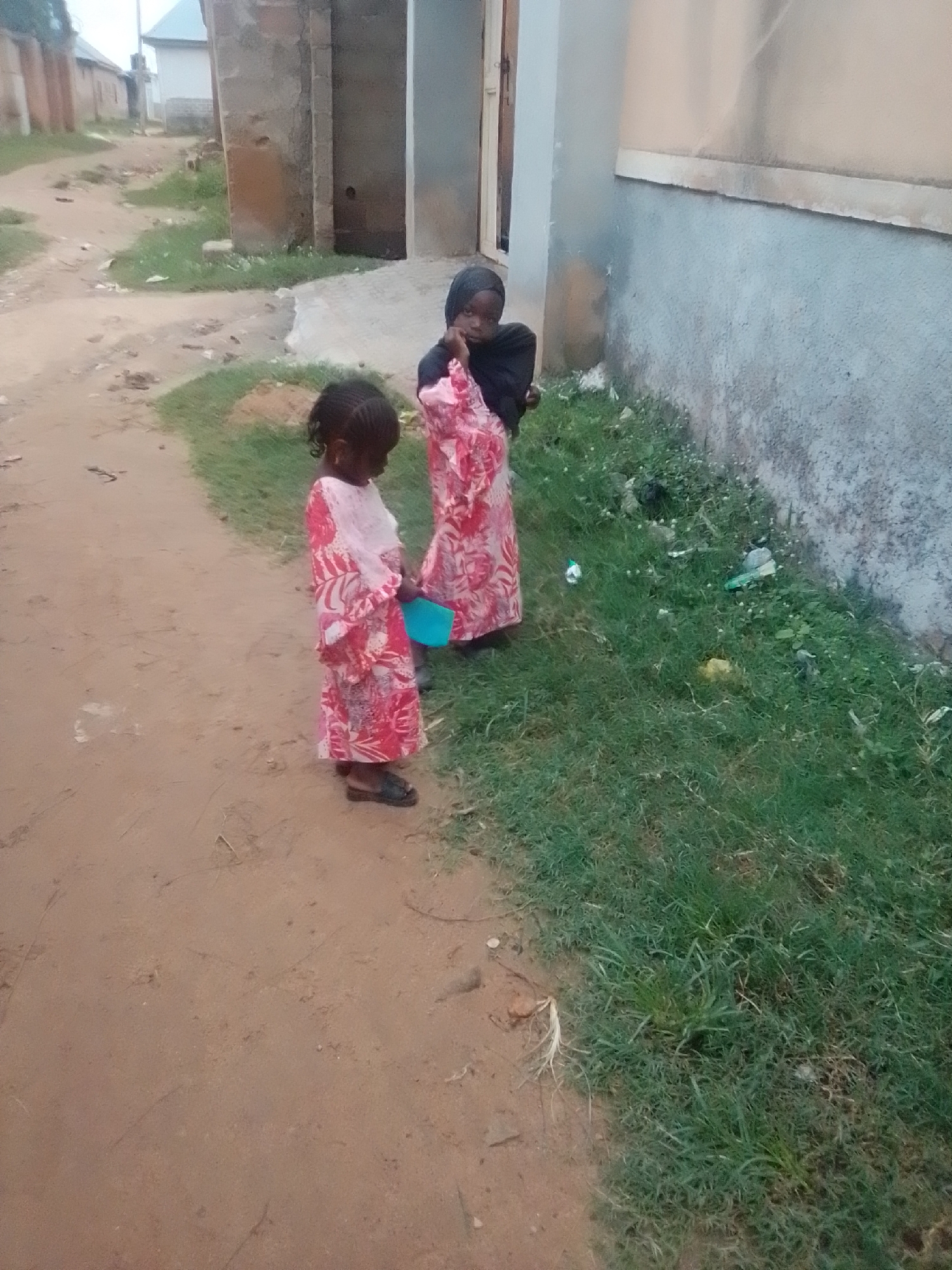 Two young children, appearing to be girls, are standing on a dirt path next to a grassy area. The child on the left is wearing a long, pink dress with a white overlay and is holding a blue bucket. The child on the right is wearing a dark head covering and a long, patterned pink garment, and is partially obscured by the other child and the building. They are standing in front of a building with a light-colored wall and a darker base. The environment suggests a rural or peri-urban setting, consistent with areas in Gombe, Nigeria, characterized by unpaved paths, modest buildings, and open grassy spaces. The scene appears to be an outdoor public space, possibly near a residential area.