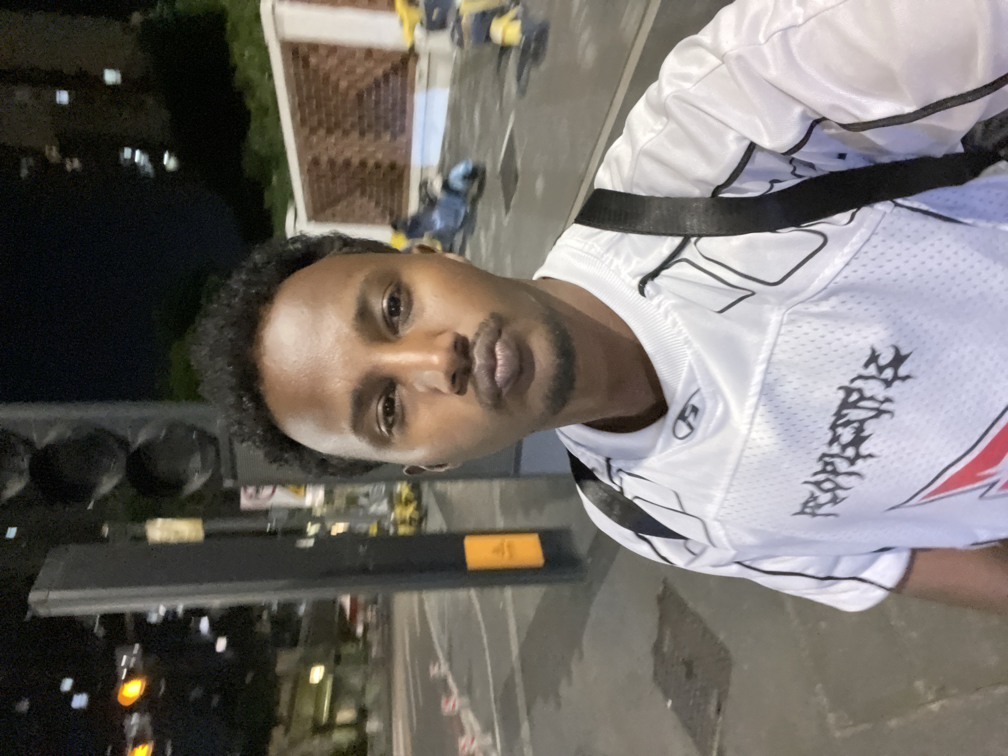 A close-up shot of a man with curly black hair and brown skin wearing a white jersey with black accents and a black strap over his shoulder. The background is a street scene at night. A traffic light is visible to the left, and a building with illuminated windows stands in the distance. The pavement is visible under the man, with road markings. The location is Chongqing High-tech Zone, China.