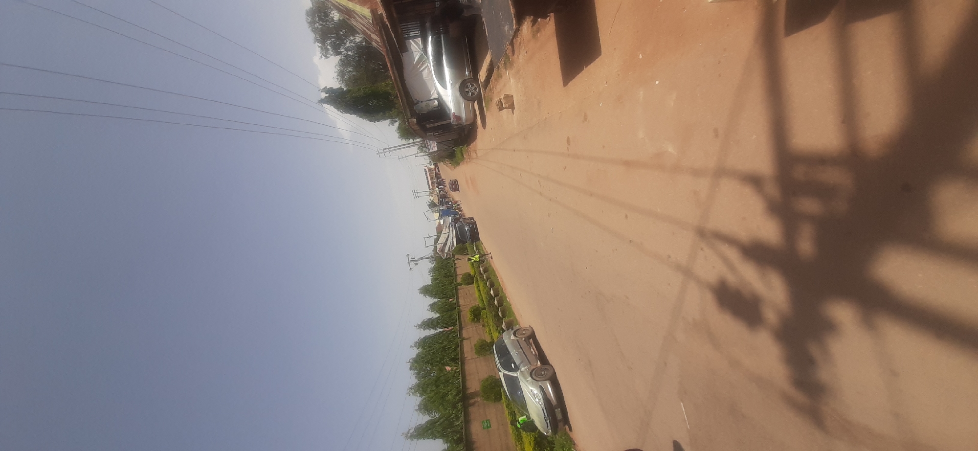 The image depicts a street scene in a seemingly ordinary town or city. The road is paved with a reddish-brown surface, possibly dirt or gravel, and has a slight incline. Several vehicles are parked or driving on the street, including a silver hatchback on the left and a silver sedan further down on the right. A dark-colored SUV is visible behind the hatchback.
A line of lush green trees runs along a low, brown wall on the left side of the street, suggesting a landscaped area or a park. On the right side, a building with a corrugated metal roof and open storefronts is visible, with some items displayed outside. Power lines crisscross the sky, supported by utility poles.
Two individuals in bright yellow vests are standing on the side of the road, possibly directing traffic or performing some sort of service. The sky is a clear, pale blue, indicating a bright, sunny day. The overall impression is a bustling but somewhat undeveloped urban environment.