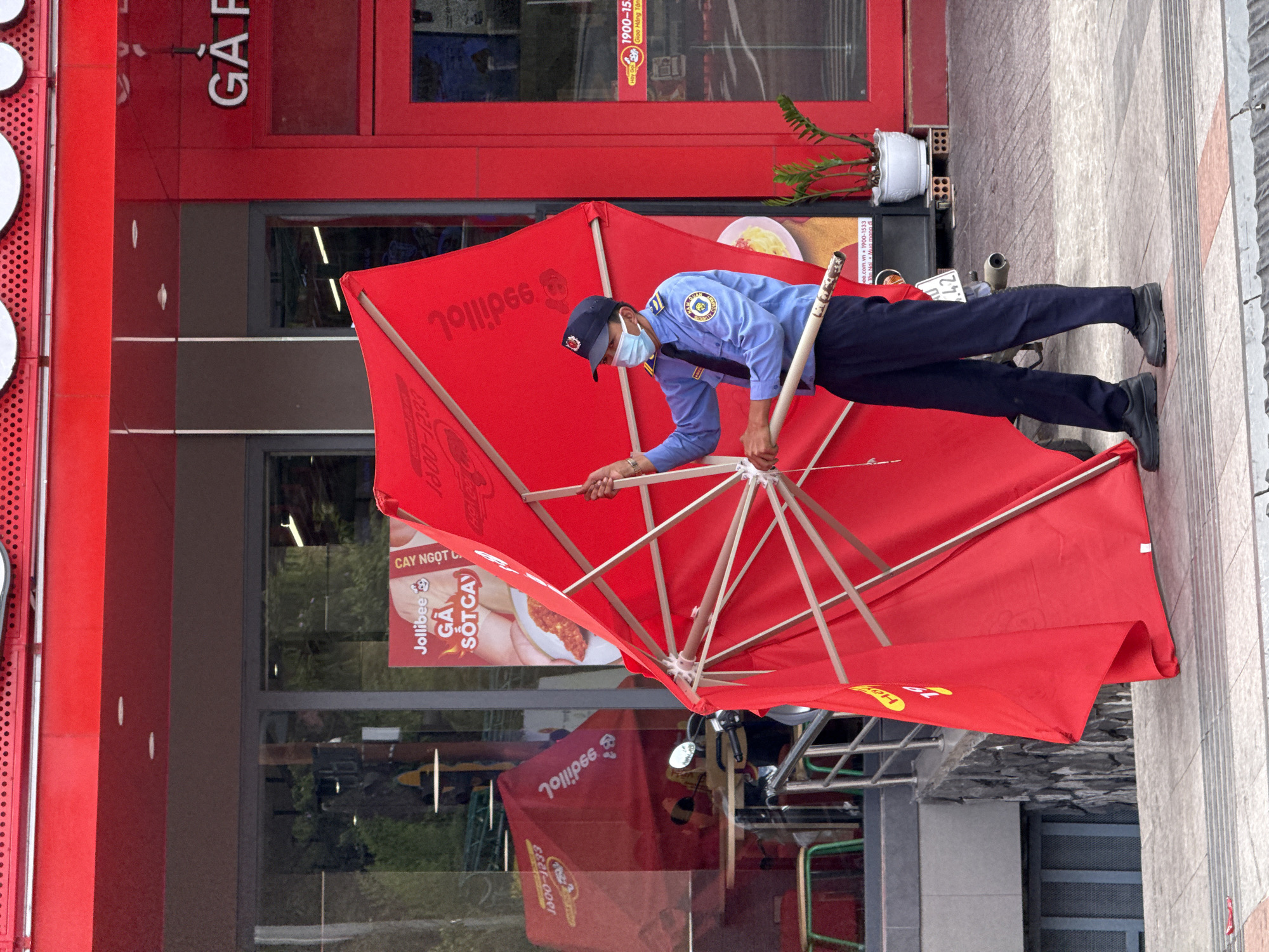 The image depicts a security guard assembling a large red Jollibee umbrella in front of a Jollibee fast-food restaurant. He is wearing a blue uniform, black pants, a black cap, and a face mask. The umbrella is open, revealing its wooden frame. The Jollibee restaurant has a red facade and large windows, and its signs are visible. There is a parked motorcycle visible behind the umbrella and a potted plant next to the restaurant entrance. The scene is located on a paved sidewalk and appears to be in an urban area, likely in Da Lat, Vietnam, based on the language in the signage.