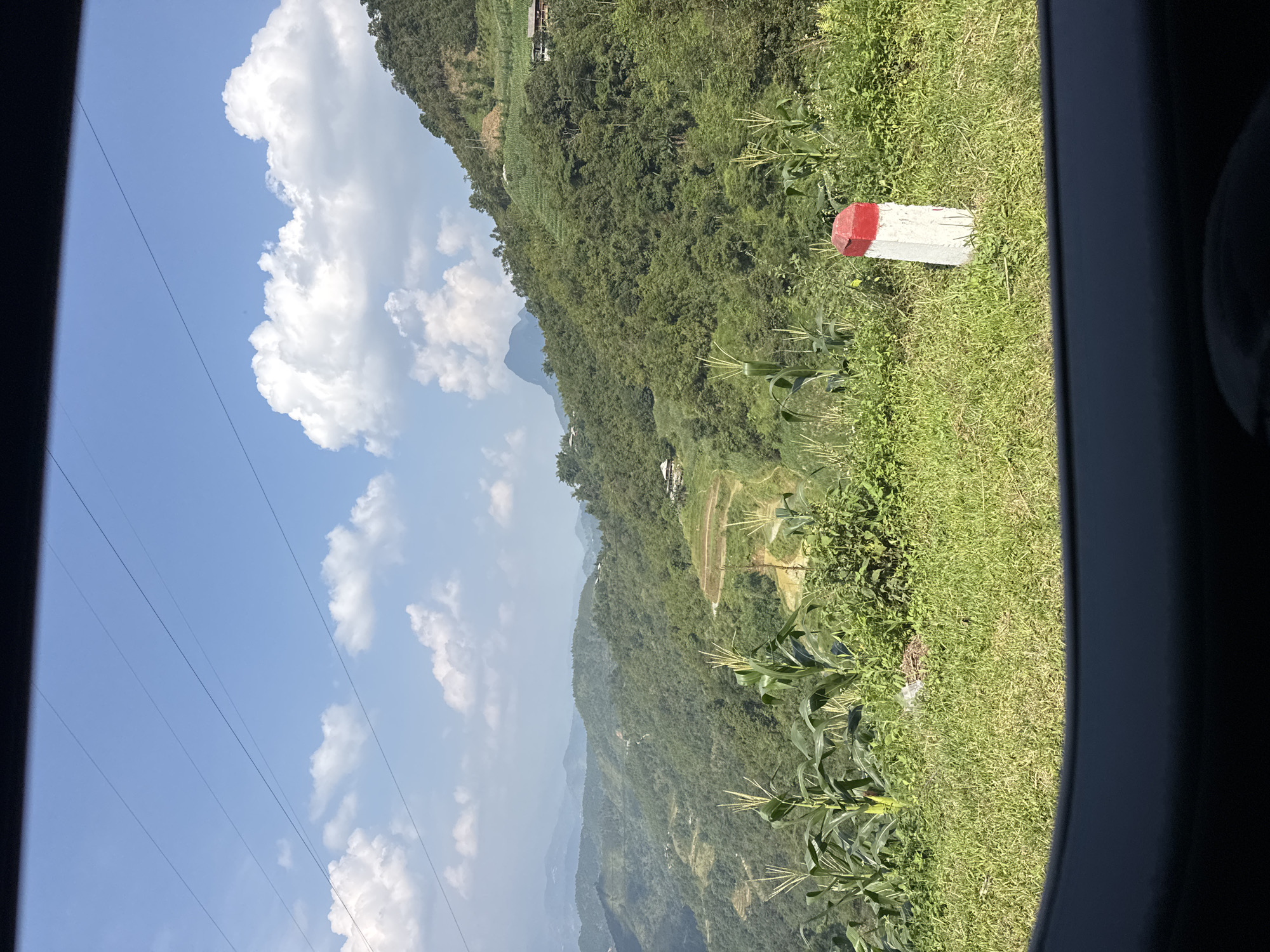 The image shows a view from inside a vehicle, likely a car, overlooking a mountainous landscape in Mậu Duệ Commune, Vietnam. The landscape consists of tiered rice paddies and vegetation-covered hills. In the foreground, there is green grass and corn plants. A white concrete marker with a red top is visible to the right of the frame. The sky is blue with scattered white clouds. Power lines are also visible against the sky.