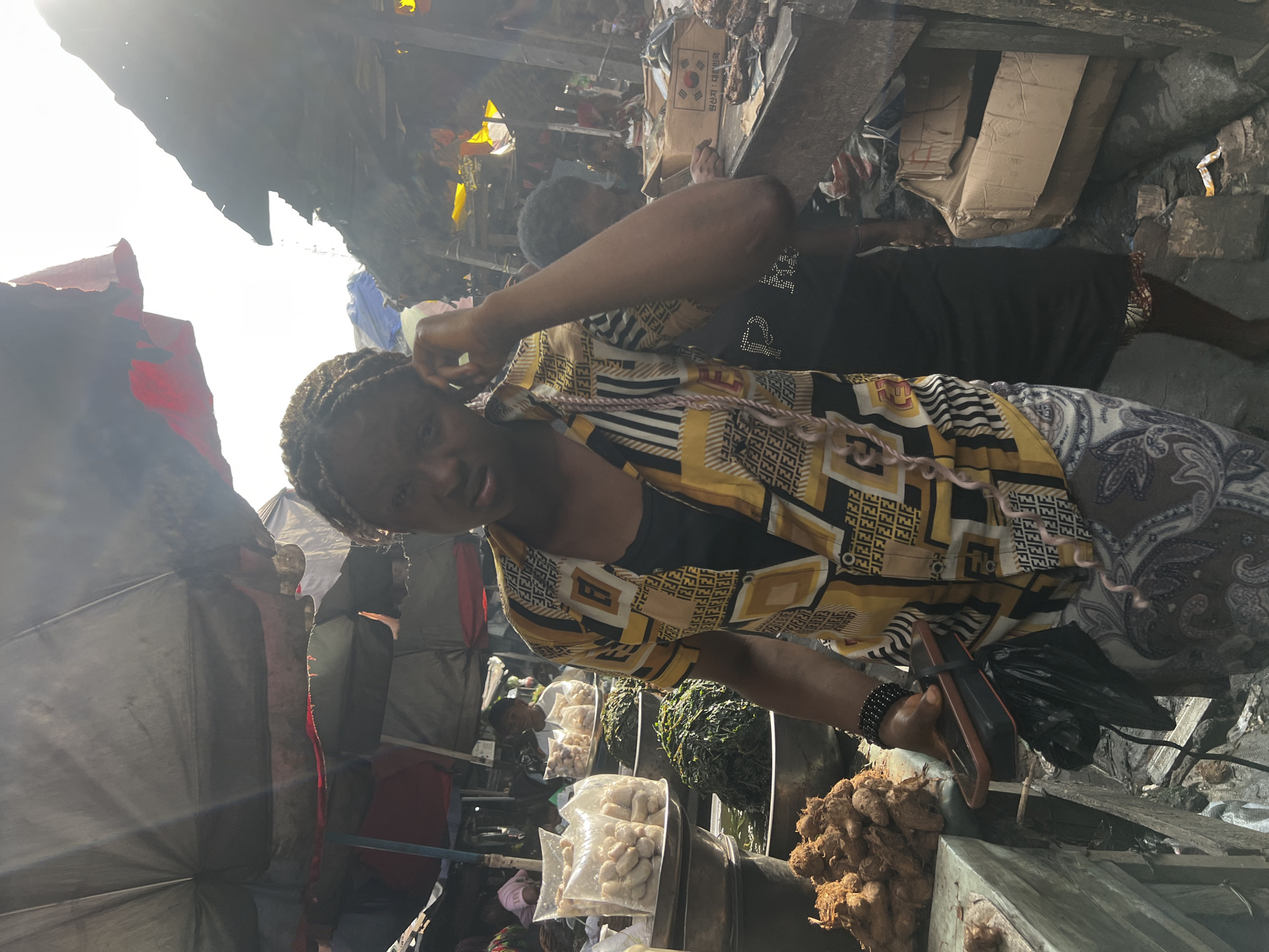 The image captures a scene at the Onitsha Main Market in Nigeria. In the foreground, a woman with braided hair is looking towards the camera, with her hand touching her head. She is wearing a yellow and black patterned shirt over a dark tank top and a patterned skirt. In her left hand, she holds a dark object, possibly a phone, with a black plastic bag attached.
The market environment is evident with various stalls and goods on display. To the left, there are plastic bags filled with small, light-colored items, likely food products, stacked on metal bowls. In front of these, a pile of brown, root-like vegetables or tubers is visible. Behind the woman in the foreground, other individuals are present, some blurred in the background, suggesting a busy market atmosphere.
Overhead, tarpaulins and umbrellas provide shade, creating a dappled lighting effect with strong sunlight filtering through. The stalls appear to be constructed with basic materials, and the ground is uneven and appears to be a mix of dirt and debris. The overall impression is of a vibrant, active marketplace.