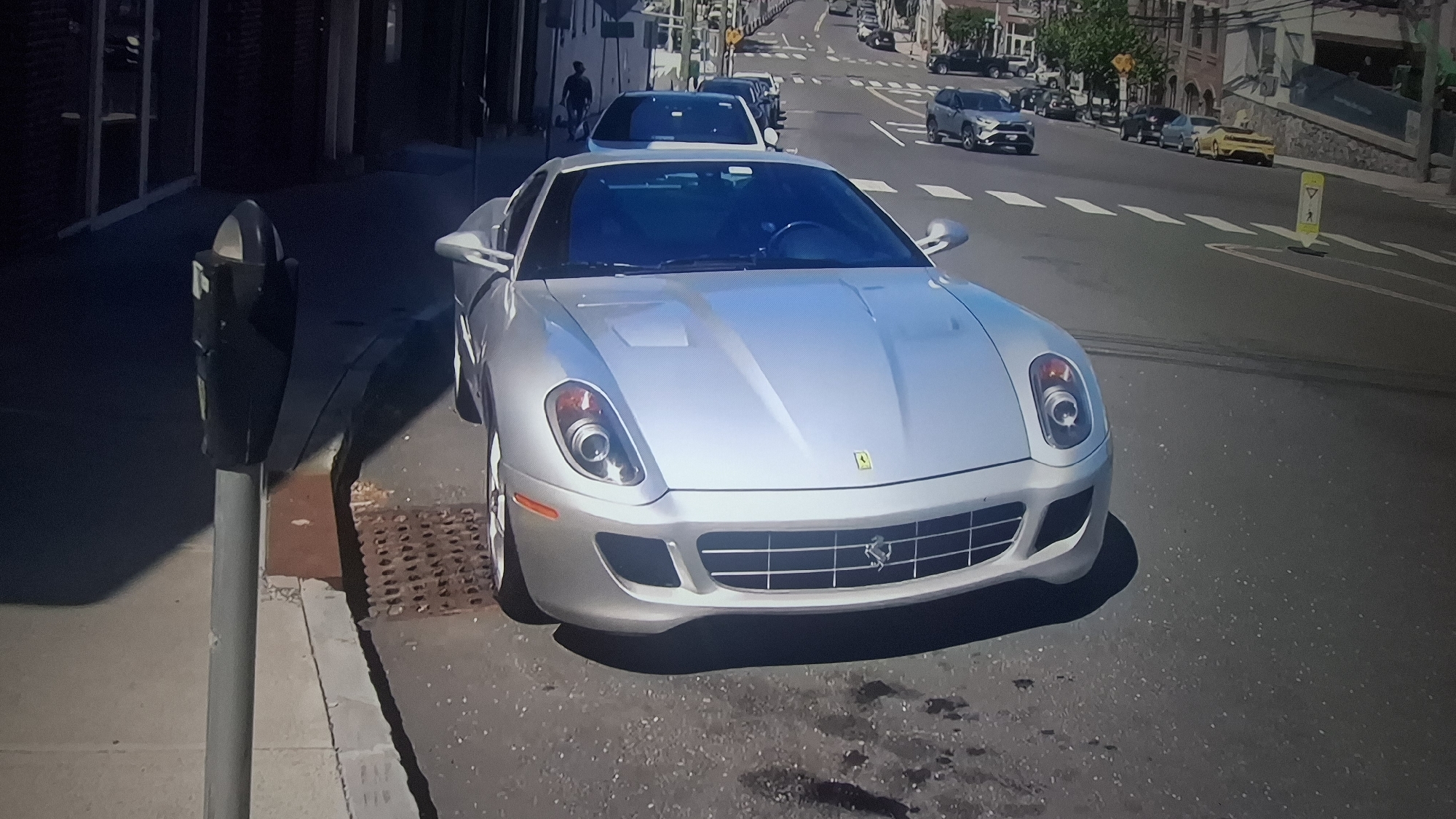 The image depicts a silver Ferrari 599 GTB parked on a city street. A parking meter stands on the sidewalk adjacent to the car. In the background, other cars are visible driving on the street, with pedestrians walking on the sidewalks. The location is an urban setting, though the exact city and state are not identifiable. Buildings line both sides of the street. The weather appears sunny.
