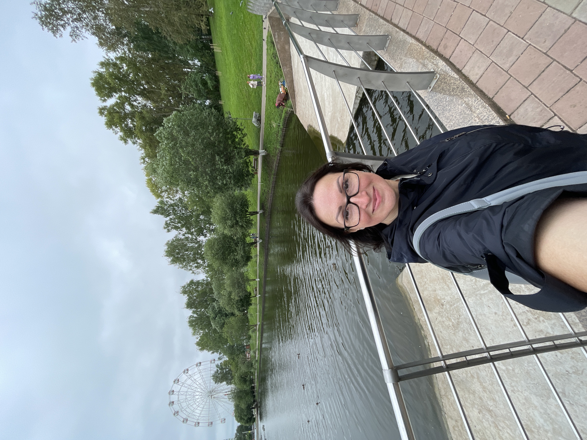 In Kirov, Russia, a woman with dark hair and glasses takes a selfie in a park. She is wearing a dark jacket and carrying a gray bag. Behind her is a body of water with ducks swimming. In the distance, a large Ferris wheel is visible, along with a tree-lined park area with people walking. The sky is overcast, suggesting a cloudy day.