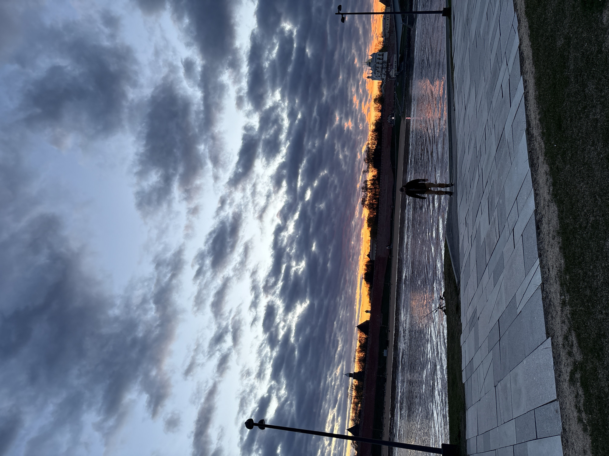 The image shows a twilight view of the Volkhov River embankment in Veliky Novgorod, Russia. The sky is filled with layered gray and white clouds. A narrow orange-lit horizon line suggests a sunset or sunrise. Along the river, the brick walls and towers of the Novgorod Kremlin are visible. A person is standing on the gray brick pavement of the embankment, close to the river's edge. The river reflects the orange light from the horizon. Street lamps line the embankment. A strip of grass separates the pavement from the river.