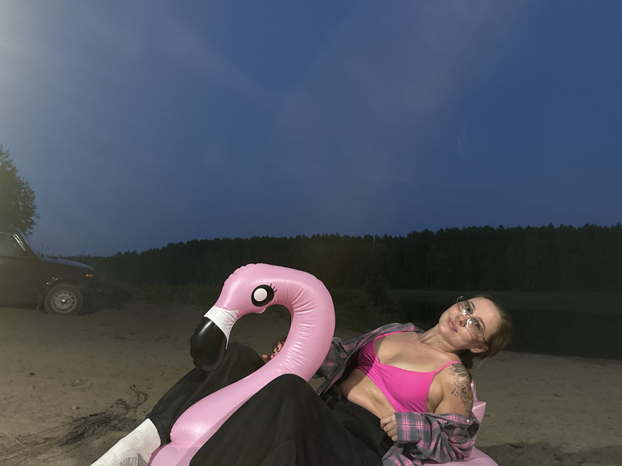 A woman with glasses and tattoos on her left arm is relaxing on an inflatable pink flamingo in a sandy area. She is wearing a bright pink bikini top and an open grey and pink plaid shirt. A dark-colored vehicle is parked to her left, and a dense line of trees forms the background. The sky above is a deep blue, indicating dusk or nighttime. The location is Garevaya, Russia.