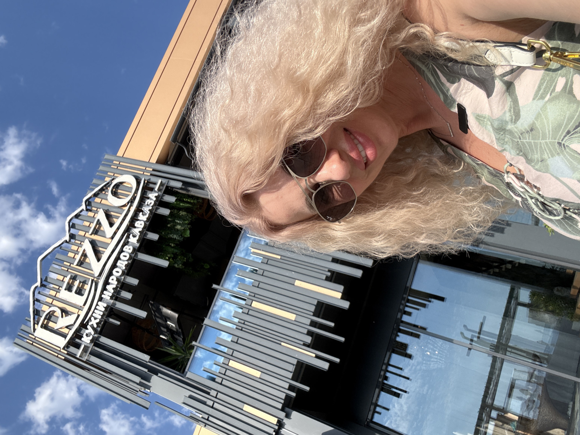 A woman with blonde, wavy hair and sunglasses smiles at the camera. She is wearing a green and white patterned dress and a silver necklace. A white purse strap with gold hardware is visible over her shoulder. She appears to be outdoors on a sunny day, with a bright blue sky and scattered clouds in the background.
Behind the woman, a modern building with large glass windows is visible. Above the windows is a sign with the text "RENZO" in large white letters, and below it, smaller text reads "КУХНИ НАРОДОВ КАВКАЗА", which translates to "CUISINE OF THE PEOPLES OF THE CAUCASUS". The building's facade features a sleek design with horizontal grey and yellow metal accents. Reflections of the sky and clouds are visible in the glass windows. The orientation of the image suggests it was taken in Sochi, Russia.