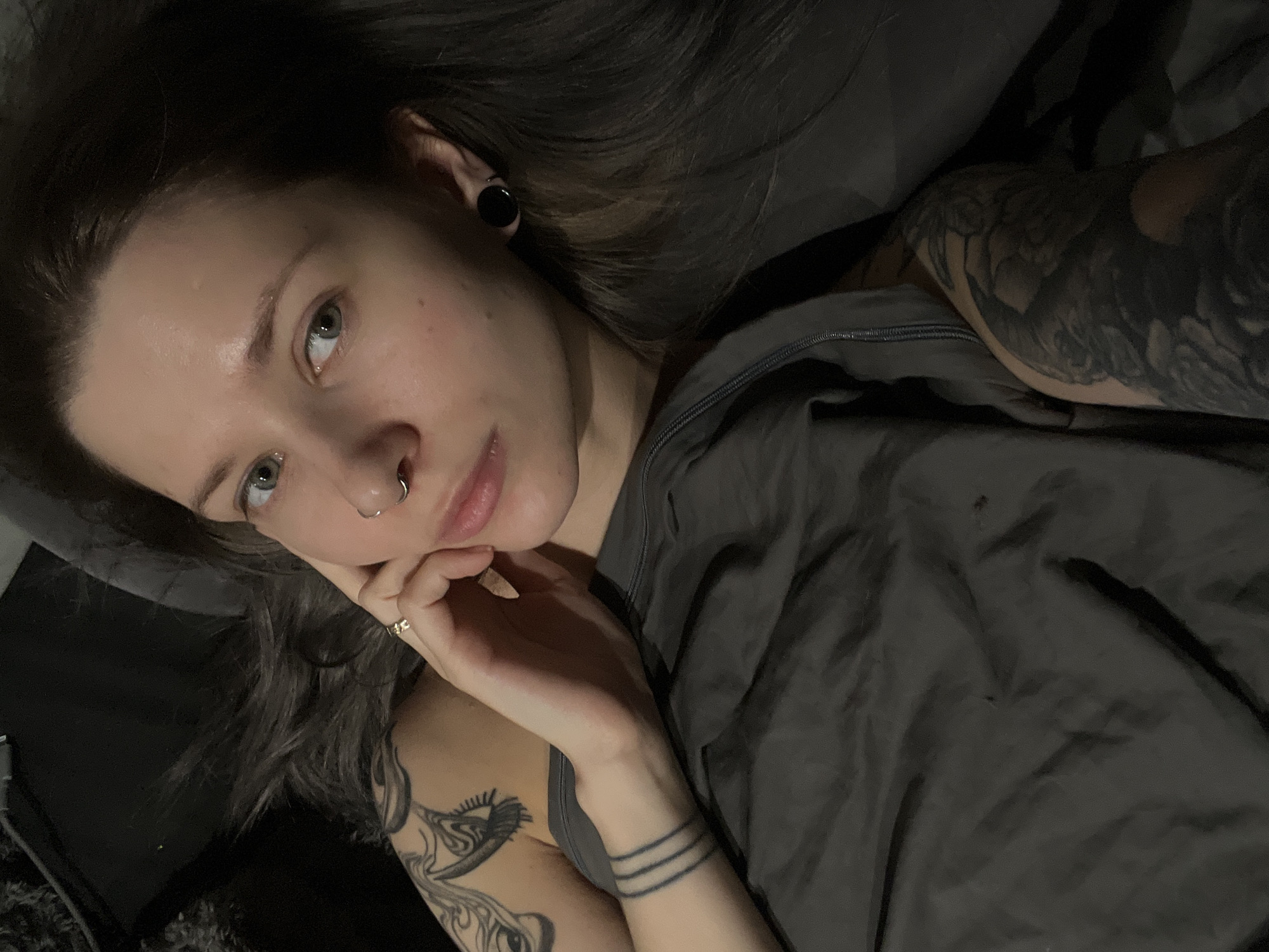 The image presents a young woman lying down indoors, captured from a slightly elevated, close-up angle. Her head is tilted, and she is looking directly into the camera with a neutral, relaxed expression.
She has long, dark hair that spills around her head, and prominent light-colored eyes, likely green or hazel. Her face features a silver septum piercing and a black ear gauge/tunnel in her left ear. She wears a grey tank top, partially visible, layered under a dark grey zipped hoodie or jacket.
Her right hand (viewer's left) is raised, with fingers gently resting against her chin and jawline. A small, subtle ring is visible on her right ring finger. Multiple tattoos are prominent: a dense, dark sleeve covers her upper left arm (viewer's right). On her right forearm (viewer's left), three distinct dark horizontal lines are tattooed around the wrist, and a partial sleeve with abstract or geometric designs extends up her bicep.
The setting appears to be a dimly lit indoor space, possibly a bedroom or living room, with dark grey fabric visible in the background, suggesting bedding or upholstery. The dim lighting creates a soft and intimate atmosphere. There is no visible text in the image.