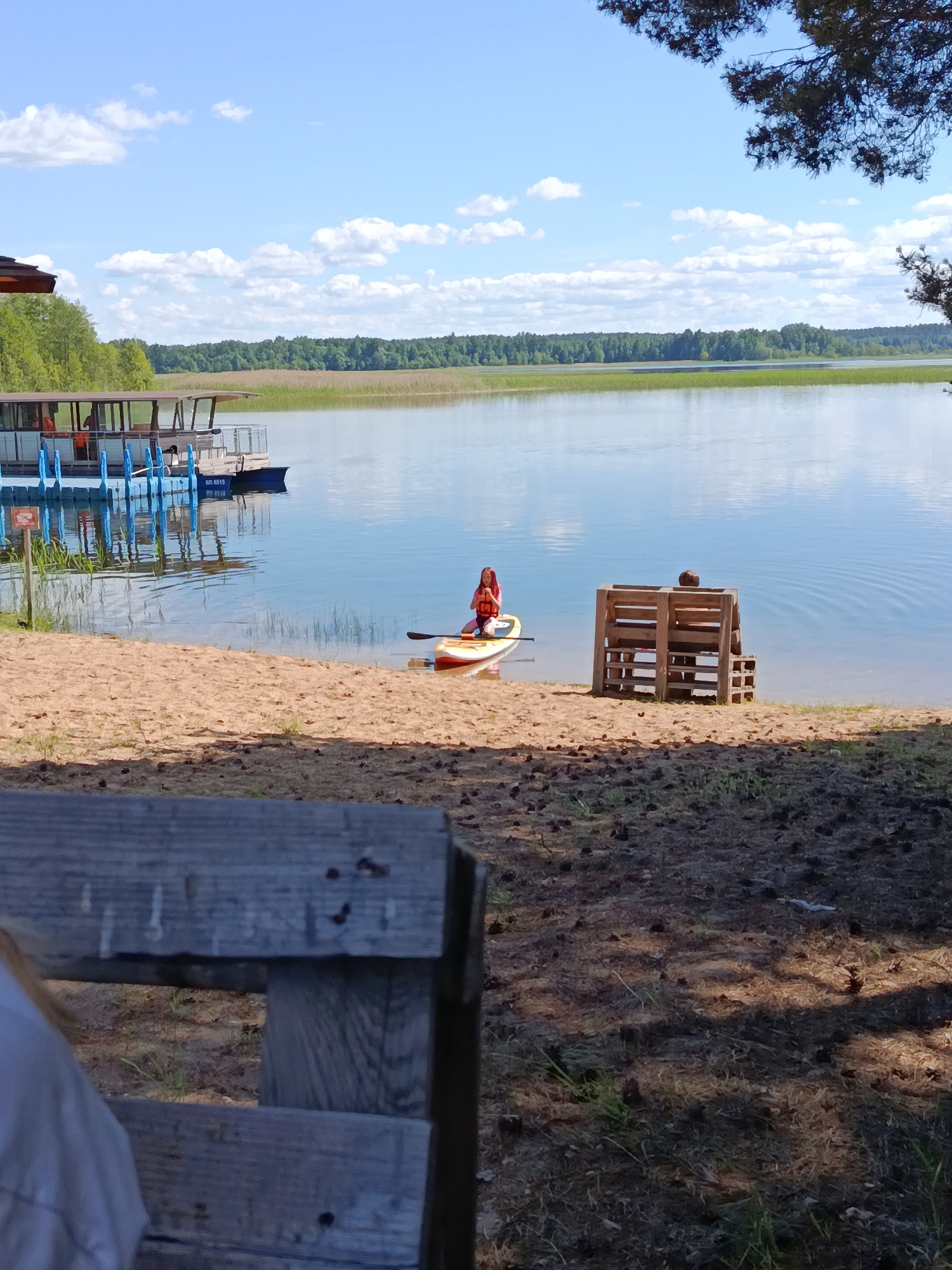 A wide shot captures a serene lake scene in Плюскі сельскі Савет, Belarus on a sunny day. The foreground is dominated by a wooden bench, partially visible, casting shadows on the sandy ground littered with pinecones. To the right, a rustic wooden bench with a backrest sits near the water's edge, occupied by a person facing the lake. In the middle ground, a young person wearing an orange life vest kneels on a paddleboard in the calm, blue water, holding a paddle. The paddleboard is positioned near the sandy shore. To the left, a floating dock with blue pilings extends from a structure that appears to be a small building with a dark roof, and a boat is tied to the dock. The background features a dense line of green trees, beyond which the sky is a clear blue with scattered white clouds. The lake reflects the sky and the surrounding greenery.