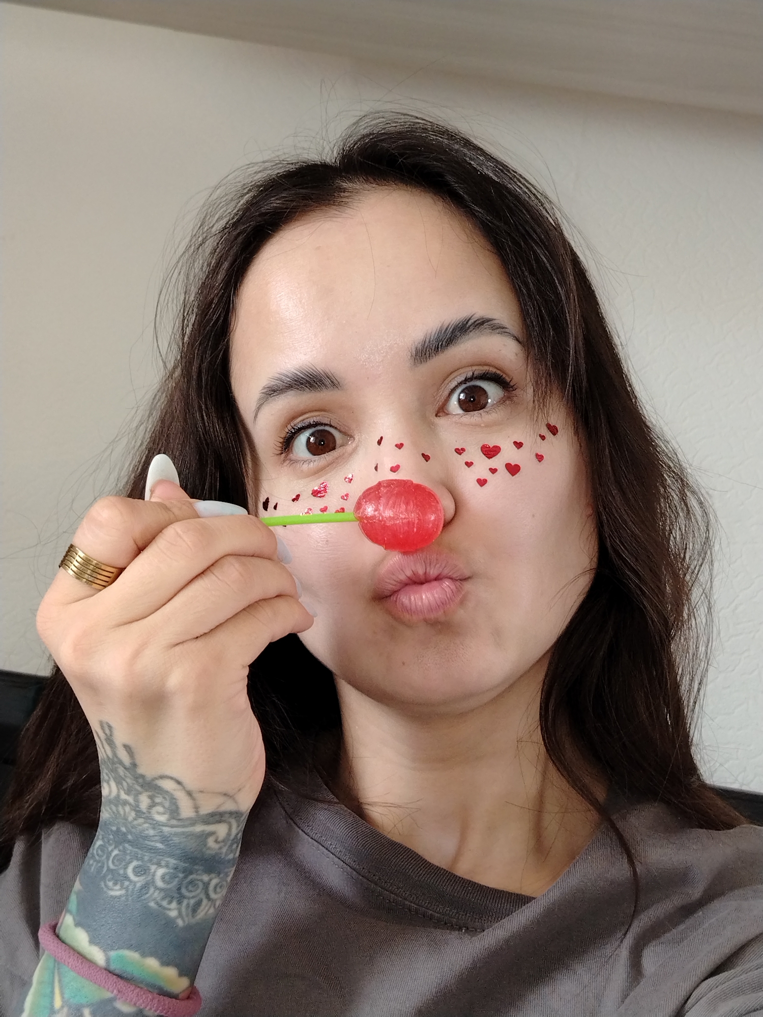A close-up, brightly lit selfie portrays a young woman indoors in Noviy, Russia, engaged in a playful activity. She has long, dark hair, brown eyes, and fair skin, with well-groomed eyebrows. Her eyes are wide open, conveying a surprised or playful expression, while her lips are puckered in a kissy face. A vibrant red, spherical lollipop on a green stick is held to her nose, resembling a clown's nose. Small, red heart-shaped stickers are scattered across both of her cheeks, adding to the whimsical theme.
The woman is wearing a casual grey t-shirt. Her left hand is prominent, holding the lollipop stick. She wears multiple stacked gold-colored rings on one finger, and her fingernails are painted white. A distinctive tattoo covers her left forearm, featuring intricate, possibly floral or mandala-like patterns in shades of grey and blue. A pink elastic hair tie is wrapped around her left wrist. The background is a plain, light-colored wall, indicating an indoor setting with natural daytime lighting. No visible text is present in the image. The overall mood is one of lightheartedness and fun.