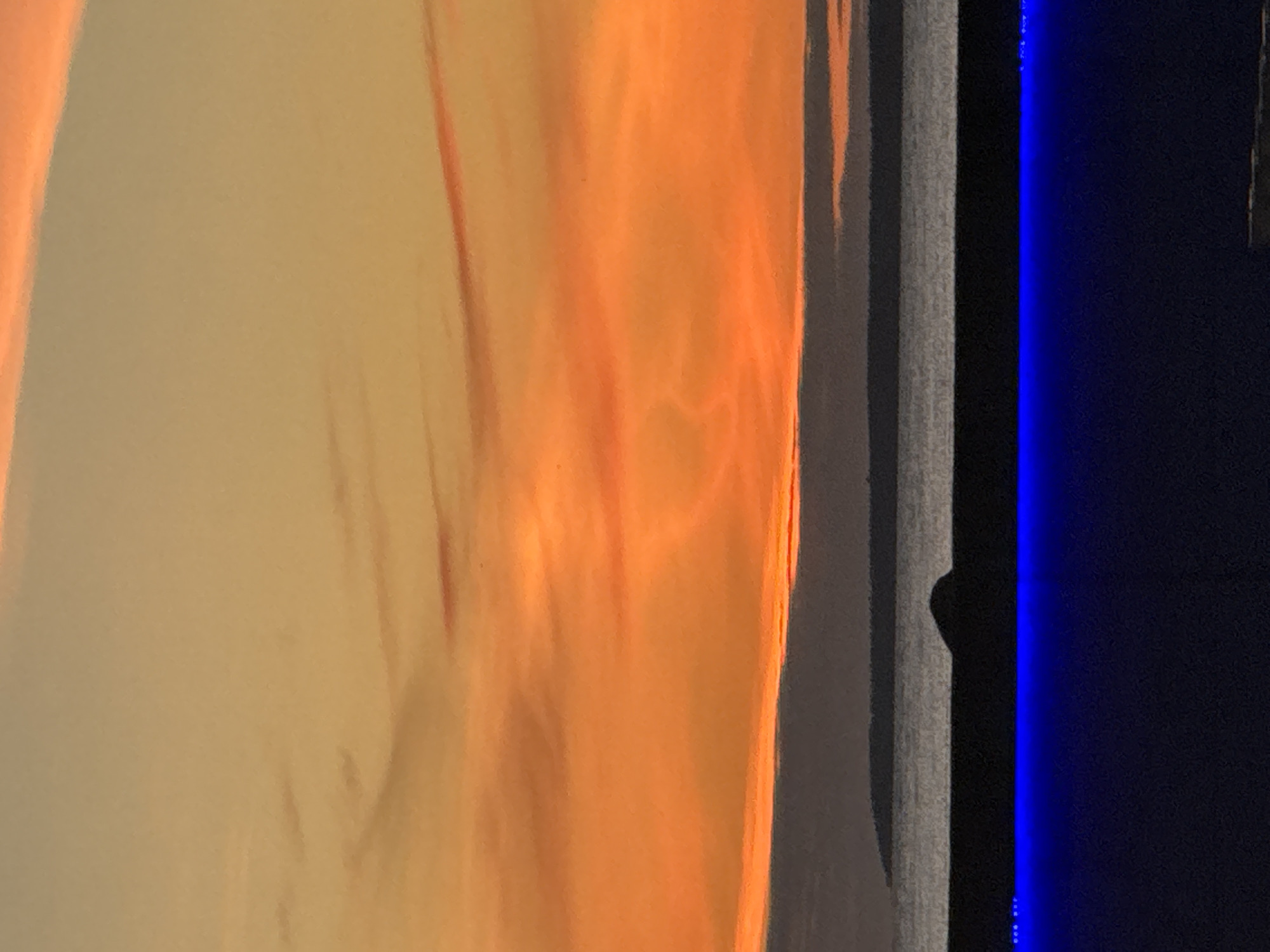 The image depicts a sunset over the Baltic Sea, viewed from a rooftop or elevated position in Tallinn, Estonia. The sky is dominated by vibrant hues of orange and yellow, with wispy clouds adding texture. In the distance, a dark silhouette of land, likely the Estonian coastline or an island, is visible against the horizon. In the foreground, a dark structure with a strip of bright blue LED lights runs along its edge, suggesting it is a modern building or amenity. The overall scene is serene and captures the natural beauty of a coastal sunset.