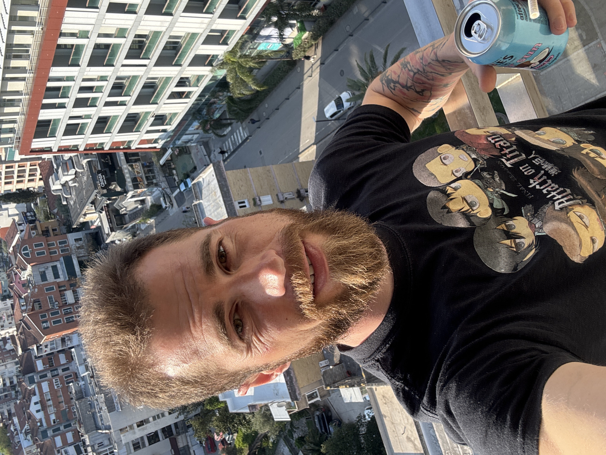 A bearded man takes a selfie from an elevated vantage point in Budva, Montenegro. He holds a can of beverage in his outstretched hand. The man's clothing includes a black T-shirt with "Attack on Titan" graphics. In the background, there is a dense urban landscape including buildings, streets, palm trees, and vehicles. A tall, white building with numerous windows is visible in the upper left. The architecture suggests a location in a built-up urban area of Budva.