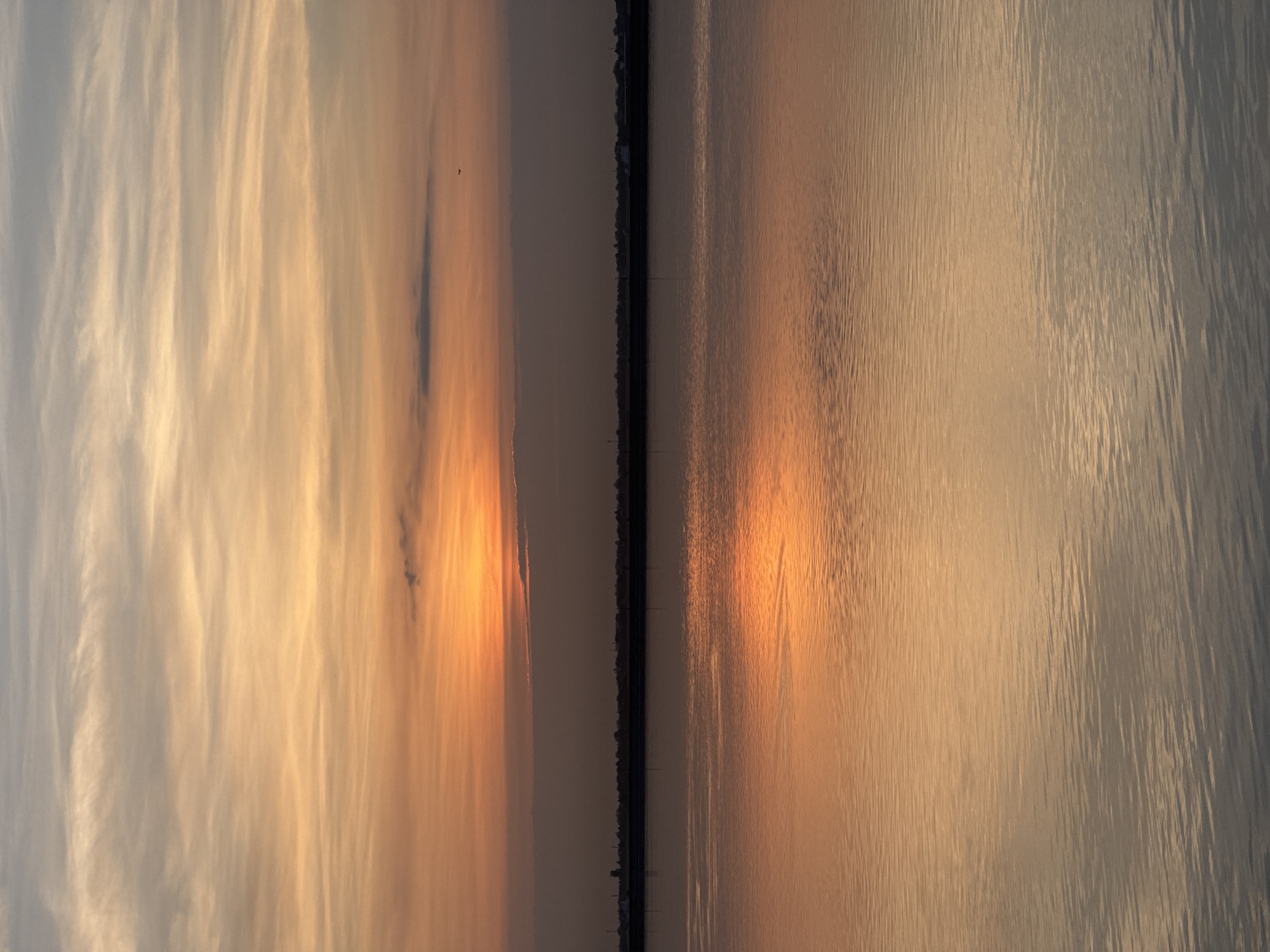 The image depicts a sunset over a body of water in Volzhsky, Russia. The sky is filled with wispy clouds illuminated with orange and yellow hues from the setting sun. This warm light is reflected in the water, creating a shimmering effect. A dark, linear structure, possibly a bridge or pier, bisects the scene horizontally.
