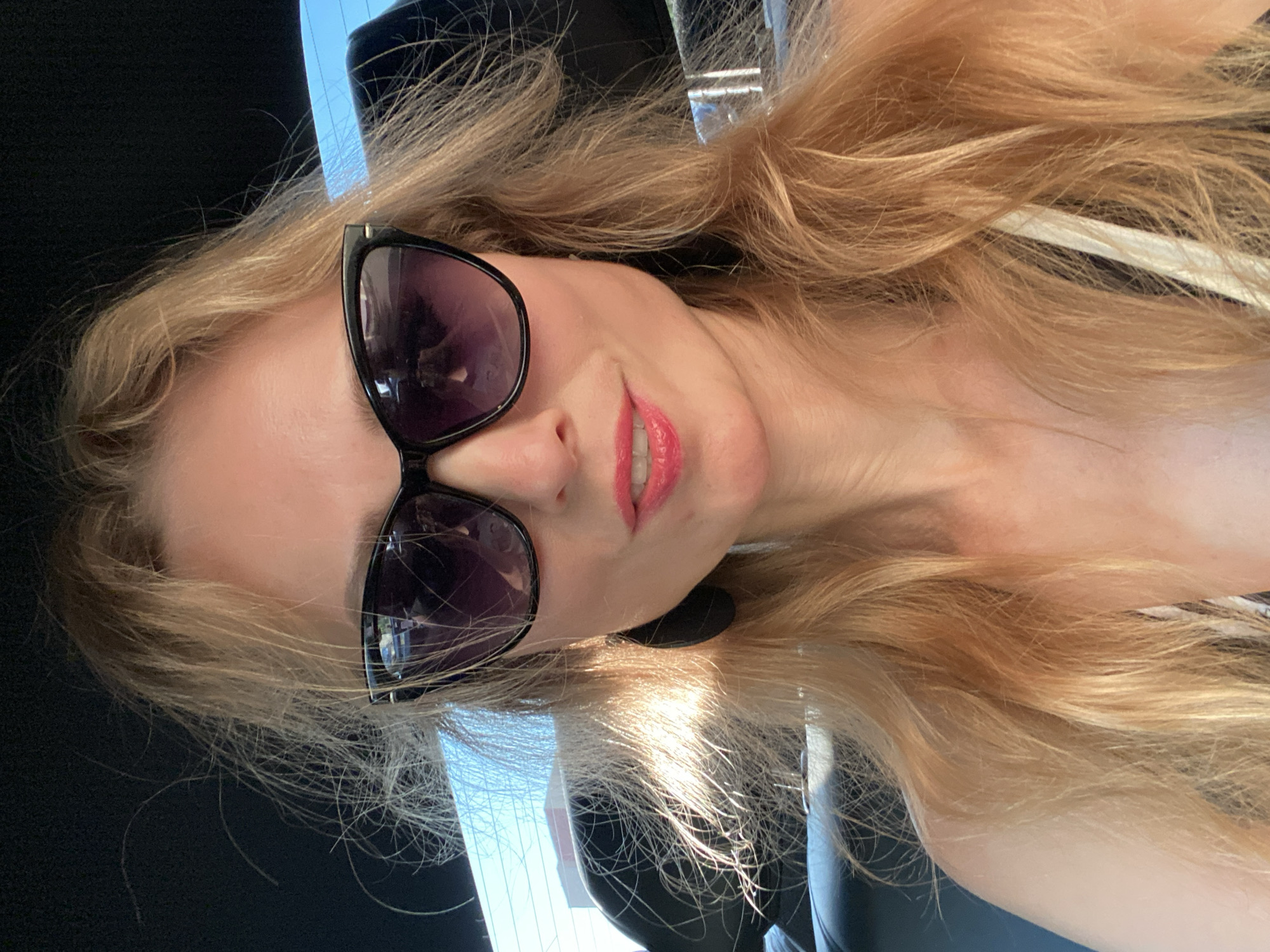 A selfie taken from a high angle shows a blonde woman in her 30s or 40s wearing black-framed sunglasses and bright pink lipstick. She is indoors, likely in a car, with the blurred background showing a car interior and a window with trees and a building visible outside. The woman's hair is a light blonde and appears windblown. She is wearing large black teardrop earrings. Her mouth is slightly open, and she appears to be smiling. The image is well-lit, with sunlight streaming in from the side, creating highlights on her hair. There are no indications of the city of Lida, Belarus, in the image.