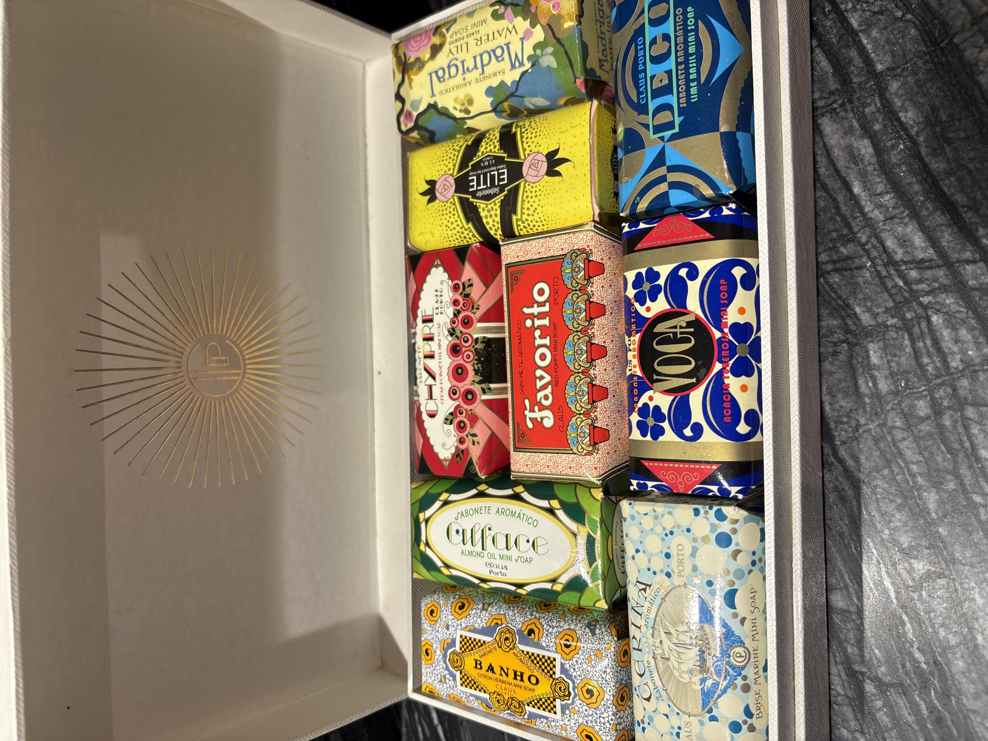 A box of assorted Claus Porto soaps is displayed on a dark, textured surface. The box itself is white with a gold embossed sunburst design on the inside lid. The soaps are arranged in two rows. The top row, from left to right, contains soaps with yellow and blue patterned wrappers, green and white wrappers, red and pink floral wrappers, bright yellow wrappers, and floral patterned wrappers. The bottom row, from left to right, has soaps with light blue and white patterned wrappers, blue and gold patterned wrappers, and blue, gold, and black patterned wrappers. The soaps are labeled with various names and scents. There are no people or animals visible in the image. The environment appears to be indoors, with no indication of the specific city of Dubai or the United Arab Emirates.