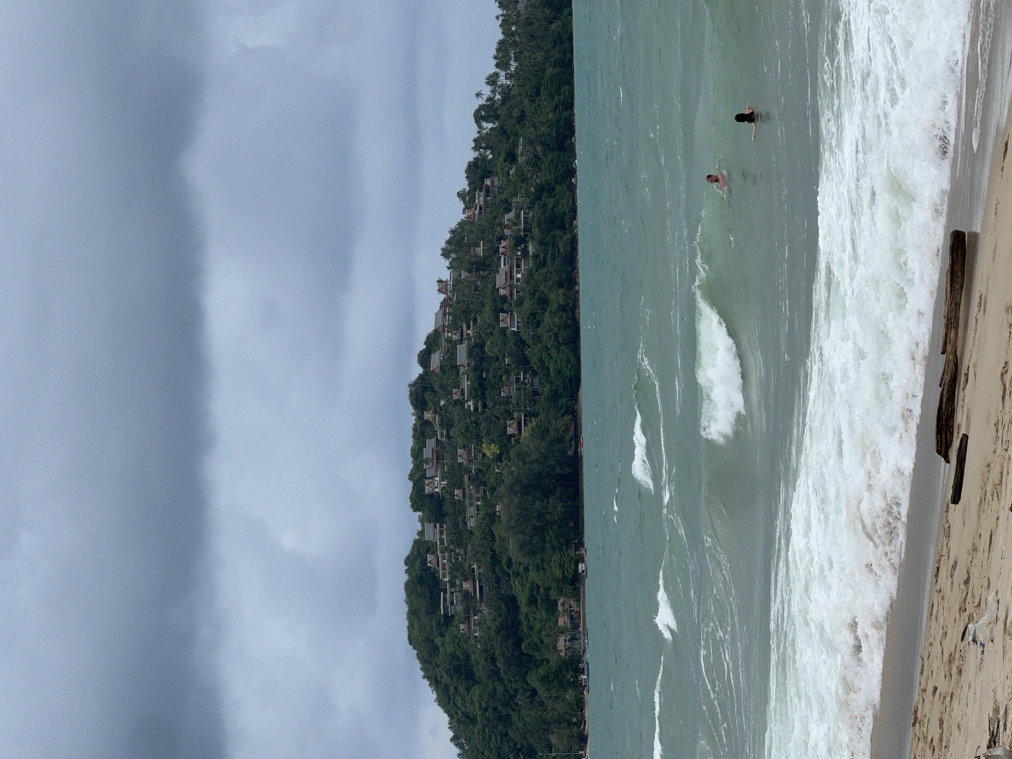 In Choeng Thale, Thailand, a sandy beach is in the foreground. Two people are swimming in the ocean. The water is a greenish-blue color with white waves crashing. In the background, a lush green hillside is covered with trees and houses, suggesting a developed area. The sky above is overcast with dark clouds. The overall scene depicts a coastal landscape with recreational activities.