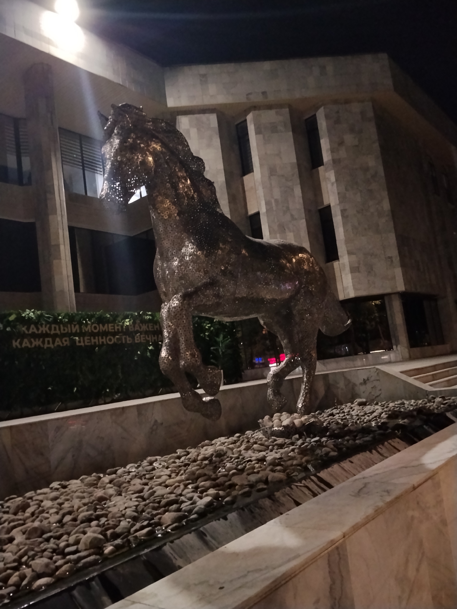 This is a night-time, low-angle, full shot of a large, dark, metallic sculpture of a horse in motion. The sculpture is made of reflective material, possibly metal plates or disks, giving it a textured, slightly fragmented appearance. The horse is in mid-stride, with its front legs extended and its head bowed slightly. The sculpture is situated in front of a modern building with a facade of light-colored stone panels and dark windows. Below the horse, there is a landscape feature with a shallow water channel filled with rounded pebbles, some of which appear wet. Behind the sculpture and in front of the building, there is a dense green hedge with golden Cyrillic lettering visible. The lettering reads: "КАЖДЫЙ МОМЕНТ ВАЖЕН" (Every moment is important) and "КАЖДАЯ ЦЕННОСТЬ ВЕЧНА" (Every value is eternal). The scene is illuminated by artificial lights, casting shadows and highlights on the sculpture and building. The image is likely taken in Bishkek, Kyrgyzstan, given the Cyrillic text.