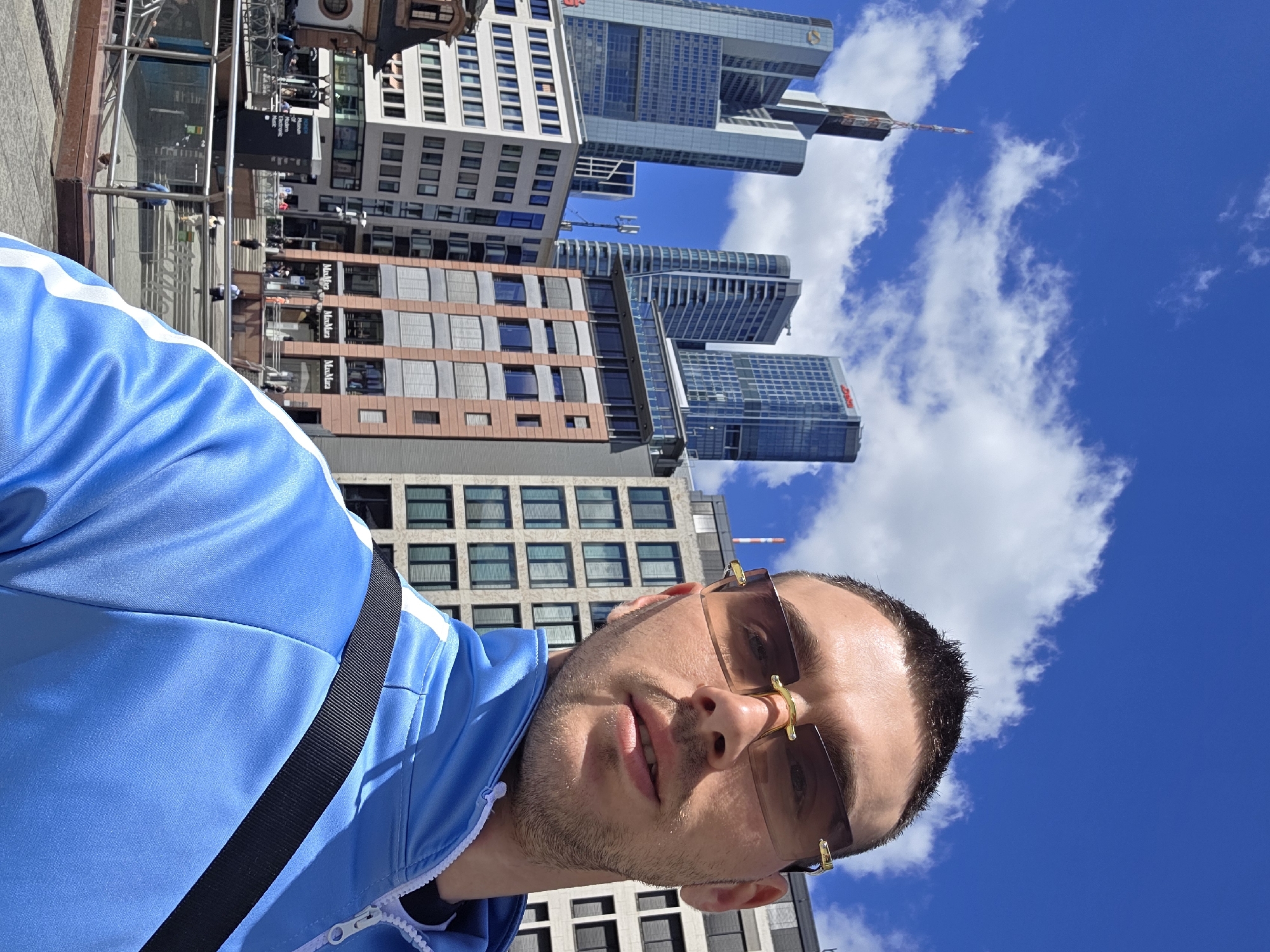 A close-up, selfie-style shot shows a man in a blue tracksuit with a black strap across his chest. He is wearing tinted rimless sunglasses. Behind him, a cityscape is visible with several modern skyscrapers and office buildings under a bright blue sky with scattered clouds. The buildings are of varying heights and architectural styles, with some featuring glass facades. The scene appears to be an urban environment, possibly a business district, given the prominence of the office buildings. The lighting suggests it is daytime with bright sunlight.