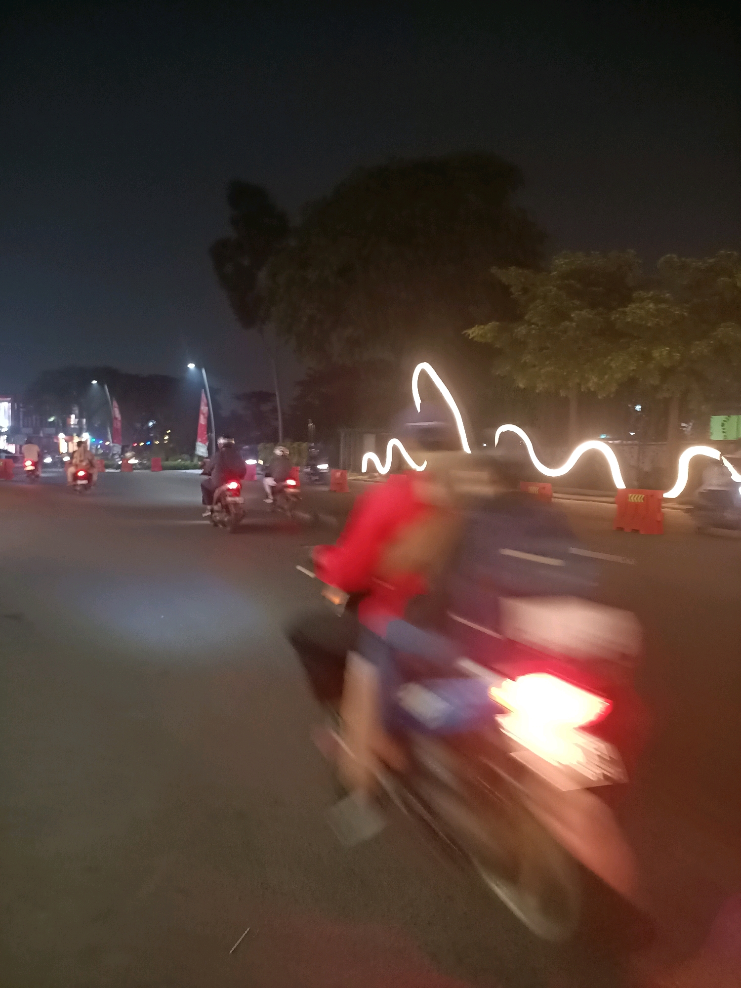 The image depicts a nighttime scene on a paved road in Telagaasih, Indonesia. A group of people are riding motorcycles, creating motion blur due to the low light and movement. The foreground shows a close-up of a motorcycle's taillight, which is brightly illuminated. In the mid-ground, several other motorcycles are visible, some with riders wearing helmets. The road is lined with orange traffic cones and trees. Decorative white string lights are also present, creating a wavy pattern in the background. The overall impression is one of activity and movement on a street at night.