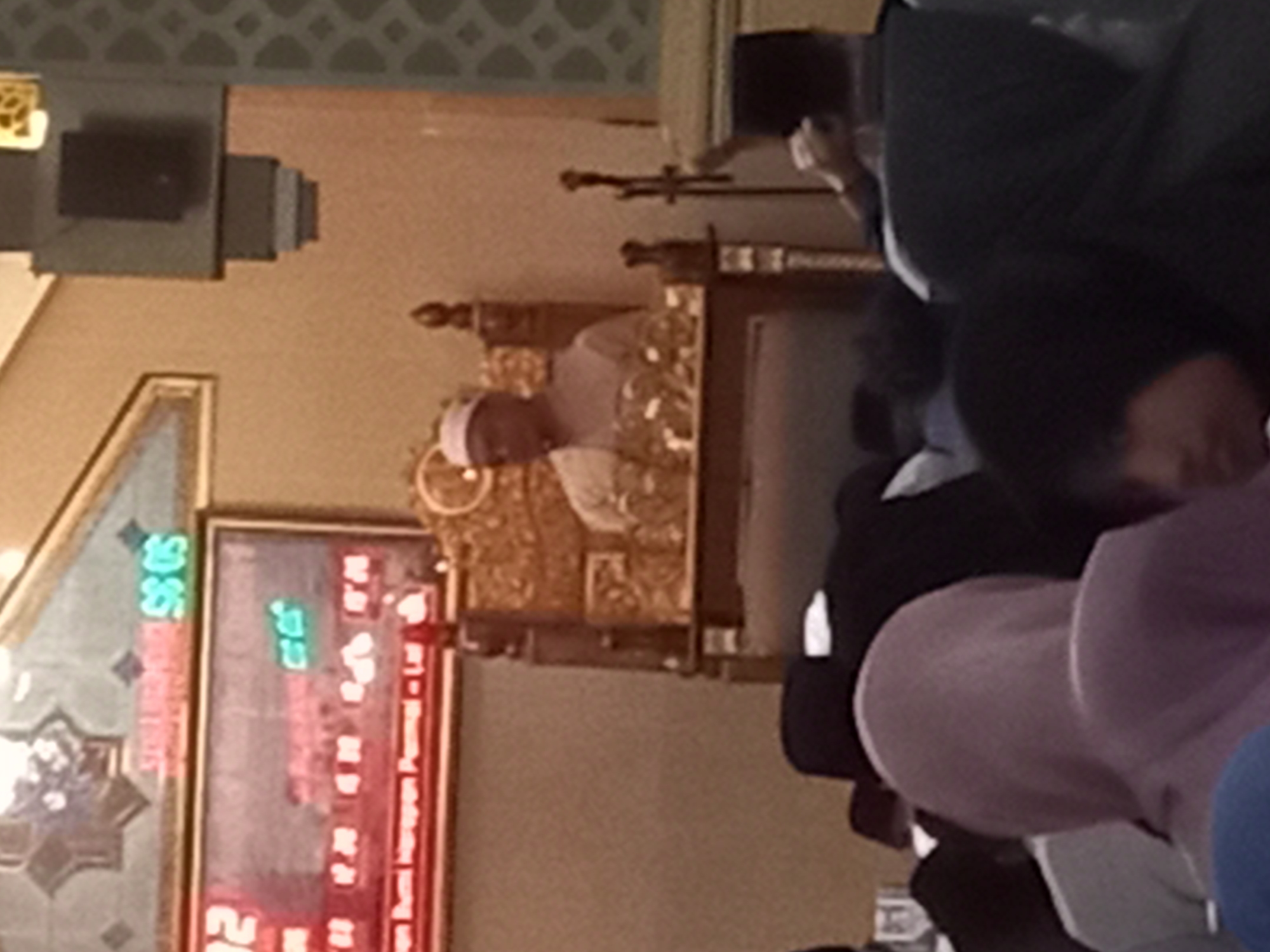 The image captures a religious gathering, likely at a mosque, in Banyumulek, Indonesia. In the background, a digital display shows numbers in red and green, indicating times or perhaps prayer schedules. Dominating the mid-ground is a man seated on an ornate, gold-colored chair. He wears a white cap and a light-colored tunic, suggesting he may be a religious leader or dignitary. The man appears to be speaking or addressing the congregation. In the foreground, several individuals are visible, facing away from the camera, indicating they are part of the audience. Their attire, including headscarves and traditional clothing, is consistent with the cultural context of Indonesia. The overall atmosphere suggests a solemn and respectful event, common in religious institutions. The lighting is ambient, with some illumination coming from the digital display and likely overhead lights, which contribute to the somewhat blurred quality of the image.