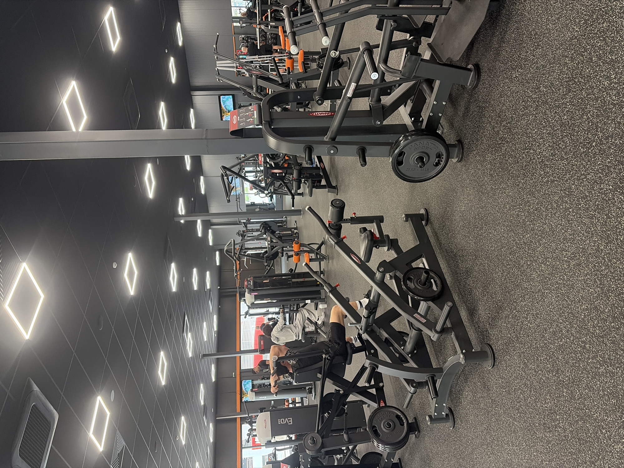 This image captures the interior of a modern gym or fitness center located in Plovdiv, Bulgaria. The setting features rows of strength training equipment, primarily plate-loaded machines, arranged on a dark grey speckled rubber floor. The ceiling is dark, adorned with numerous illuminated rectangular and diamond-shaped LED lights, creating a distinctive pattern.
Several individuals, mostly men, are present and engaged in exercise. In the mid-ground, towards the right, a person is seen operating a chest press machine, lifting weights. Further in the background, other gym-goers are visible utilizing various pieces of equipment, including what appears to be rowing machines and other selectorized or plate-loaded stations. People are dressed in typical gym attire, such as shorts, t-shirts, and athletic shoes.
Notable objects include black and orange weight machines from brands like "PANATTA" and "EVO!". Many weight plates are visible on the machines, with some clearly marked "20 KG - 44 LB". Small television screens are mounted on walls in the background, though the content displayed is not clearly discernible. The overall atmosphere suggests a well-maintained and busy fitness facility, focused on strength and resistance training. The artificial lighting indicates an indoor scene, making the time of day indeterminate. No specific weather conditions are relevant.