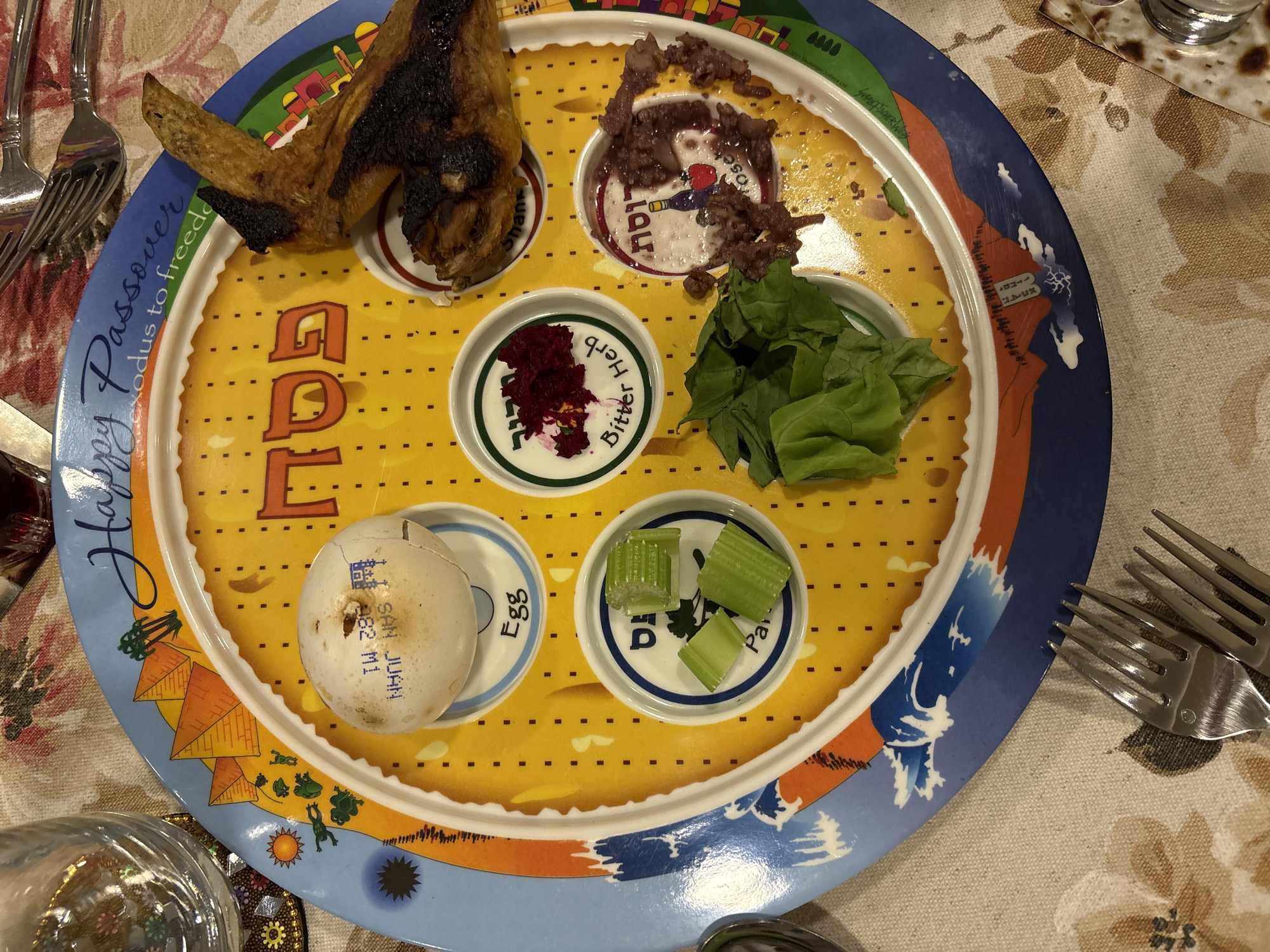 The image shows a Seder plate, used during the Jewish holiday of Passover, positioned on a tablecloth. The plate is divided into sections, each containing symbolic food items: a roasted shank bone, a mixture of apples, nuts, and wine (charoset), bitter herbs, lettuce, celery, and a hard-boiled egg. The plate has the word "Pesach" written on it in Hebrew letters, and its perimeter is decorated with scenes depicting the Exodus story. Forks are visible on both sides of the plate. The city of Huixquilucan de Degollado, Mexico is not identifiable.