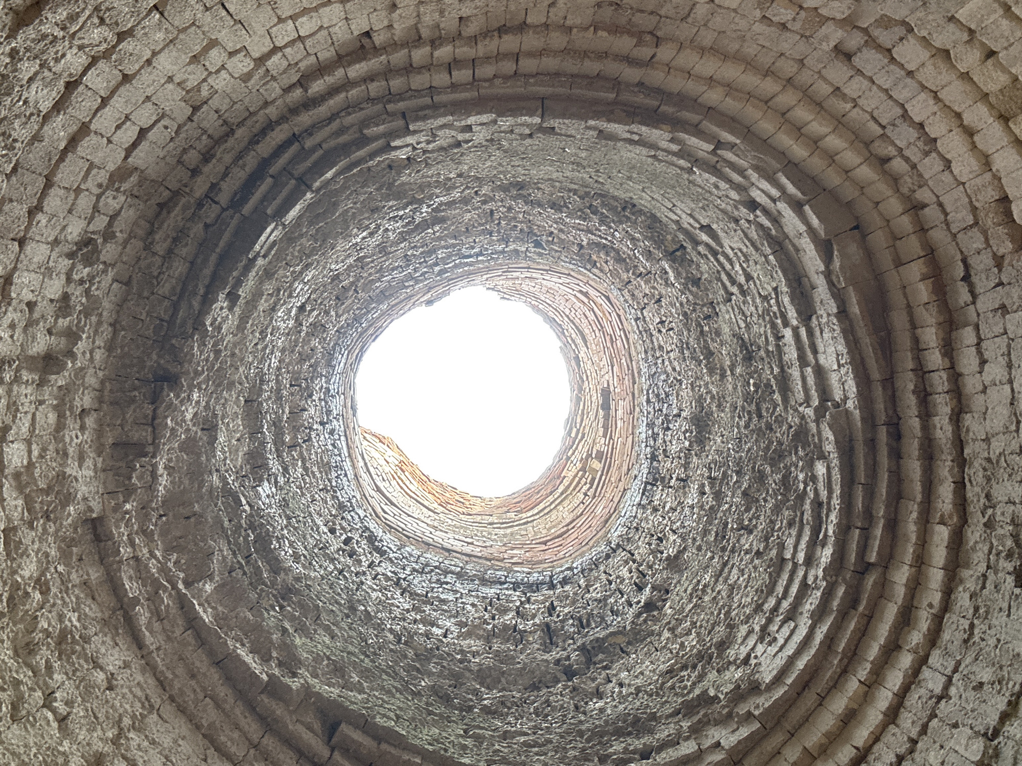 This media file is an upward-facing, wide-angle shot of the interior of a round, brick structure, likely a well or chimney shaft, in Mezhevoy, Russia. The rough, aged bricks are arranged in concentric circles, forming a tunnel-like effect that narrows towards a bright, circular opening at the top. The texture of the bricks varies, with some appearing smoother and more uniform, while others are rough and irregular, suggesting wear and tear over time. The light streaming from the opening is blown out, indicating it might be daytime. The scene evokes a sense of depth and enclosure, with the weathered brickwork hinting at the structure's age and potential historical significance. No people, text, or specific weather conditions are visible, but the overall impression is one of an old, possibly abandoned, industrial or utilitarian structure.
