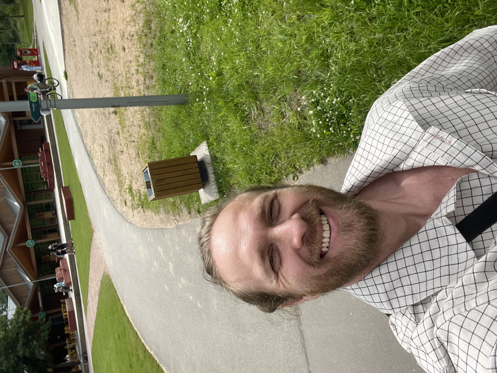 This media file is a selfie taken outdoors on a bright, sunny day, suggesting midday or early afternoon. The subject is a man with a beard and blond hair, smiling broadly at the camera. He is wearing a white checkered shirt.
The setting appears to be a park or outdoor recreation area. In the background, there's a paved walkway winding through a grassy area dotted with small white flowers. To the left, a modern building with large windows and wooden accents is visible, possibly a visitor center or cafe, with outdoor seating and tables. Further in the background, people can be seen walking and cycling. A signpost with green directional signs is also present. A wooden trash receptacle is next to the walkway.
The overall mood is cheerful and relaxed, with the subject's expression conveying happiness. The lush green grass and bright sunlight indicate pleasant weather. While the exact location is not explicitly stated in the image itself, the provided context identifies it as Sosnoviy Bor, Russia. There is no visible text in the image that provides further information about the event or specific location.
