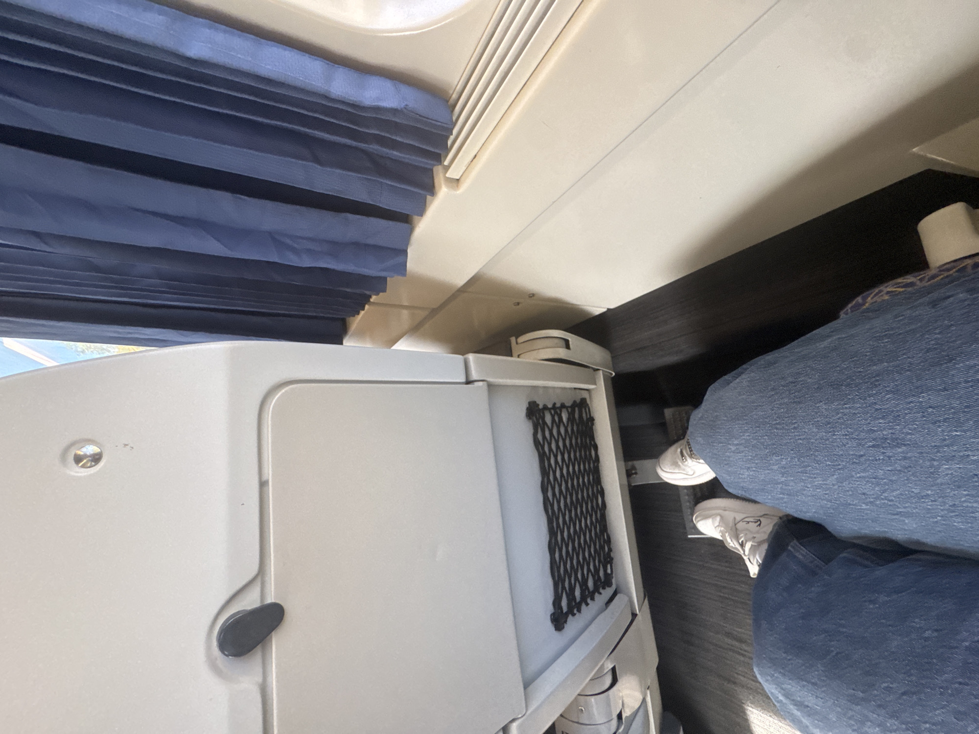 The image depicts an interior view, possibly of a train or airplane, with a blue pleated window shade partially visible at the top left. A white storage compartment or cabinet with a black mesh pocket on its side is situated on the left. The feet and lower legs of a person wearing blue jeans and white sneakers are visible in the bottom right, suggesting someone is seated in the space. The scene provides no information that can be used to deduce if the photo was taken in Trofa, Portugal.