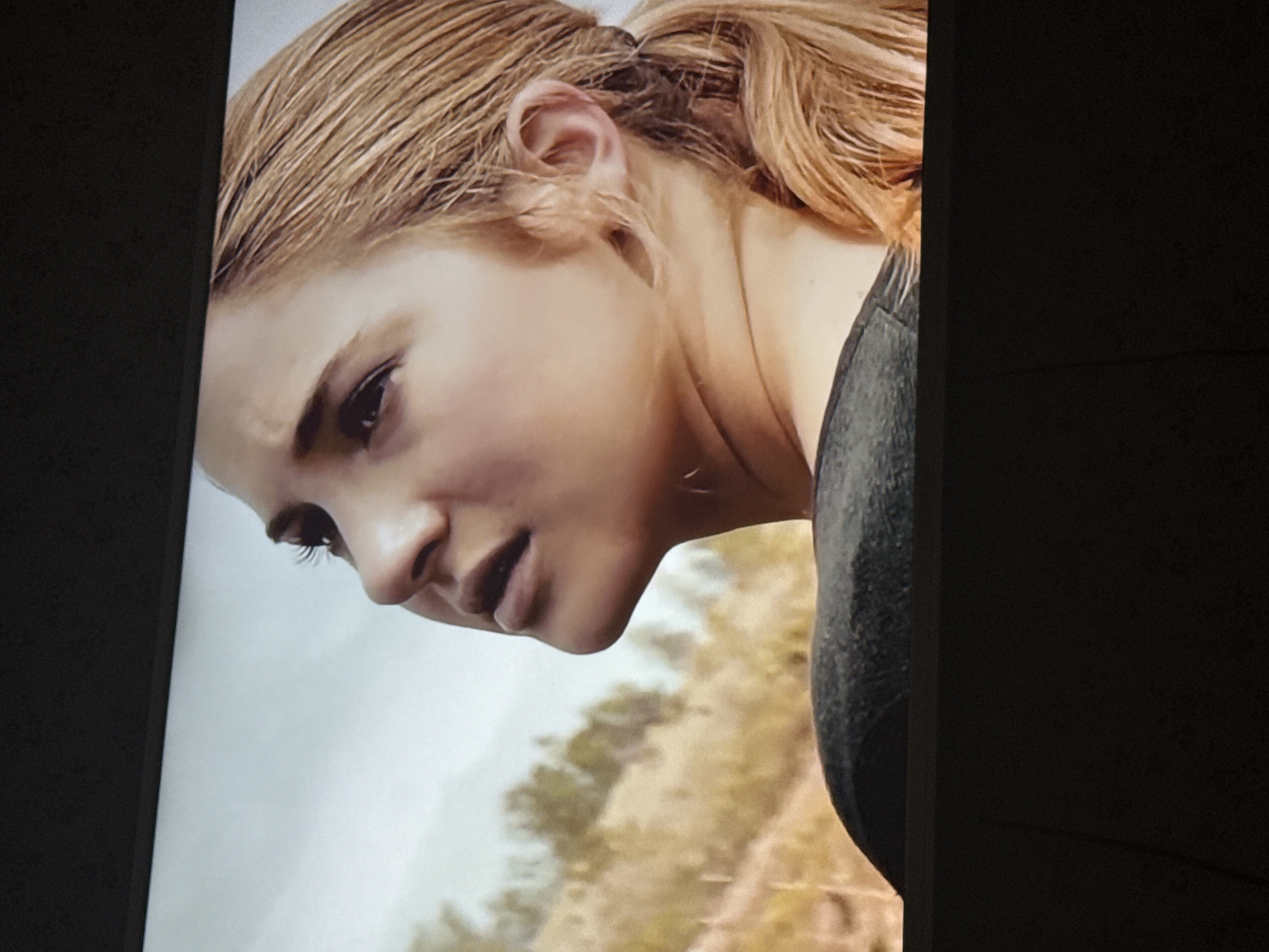 A close-up shot shows the face of a woman with light brown hair pulled back into a ponytail. She is looking down and to the left, with a furrowed brow. Her expression suggests concentration or concern. She is wearing a dark-colored garment. The background is blurred, suggesting an outdoor scene with trees or foliage. The city of Mytishchi, Russia, cannot be determined from this image.