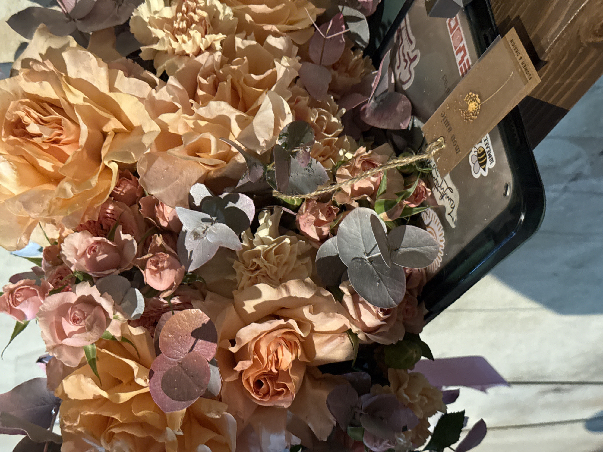 The image shows a floral arrangement in a dark container, likely a gift. The arrangement includes peach and pink roses, carnations, and grey-green eucalyptus leaves. A small tag is attached with twine, bearing a logo and the words "mon amie" and "flower & design." A sticker with a bee image is also visible on the container. The arrangement appears to be placed indoors, possibly on a white-painted wooden surface. There is no explicit indication of the location being Yerevan, Armenia, unless there is specific knowledge linking the "mon amie" flower shop logo to that city.
