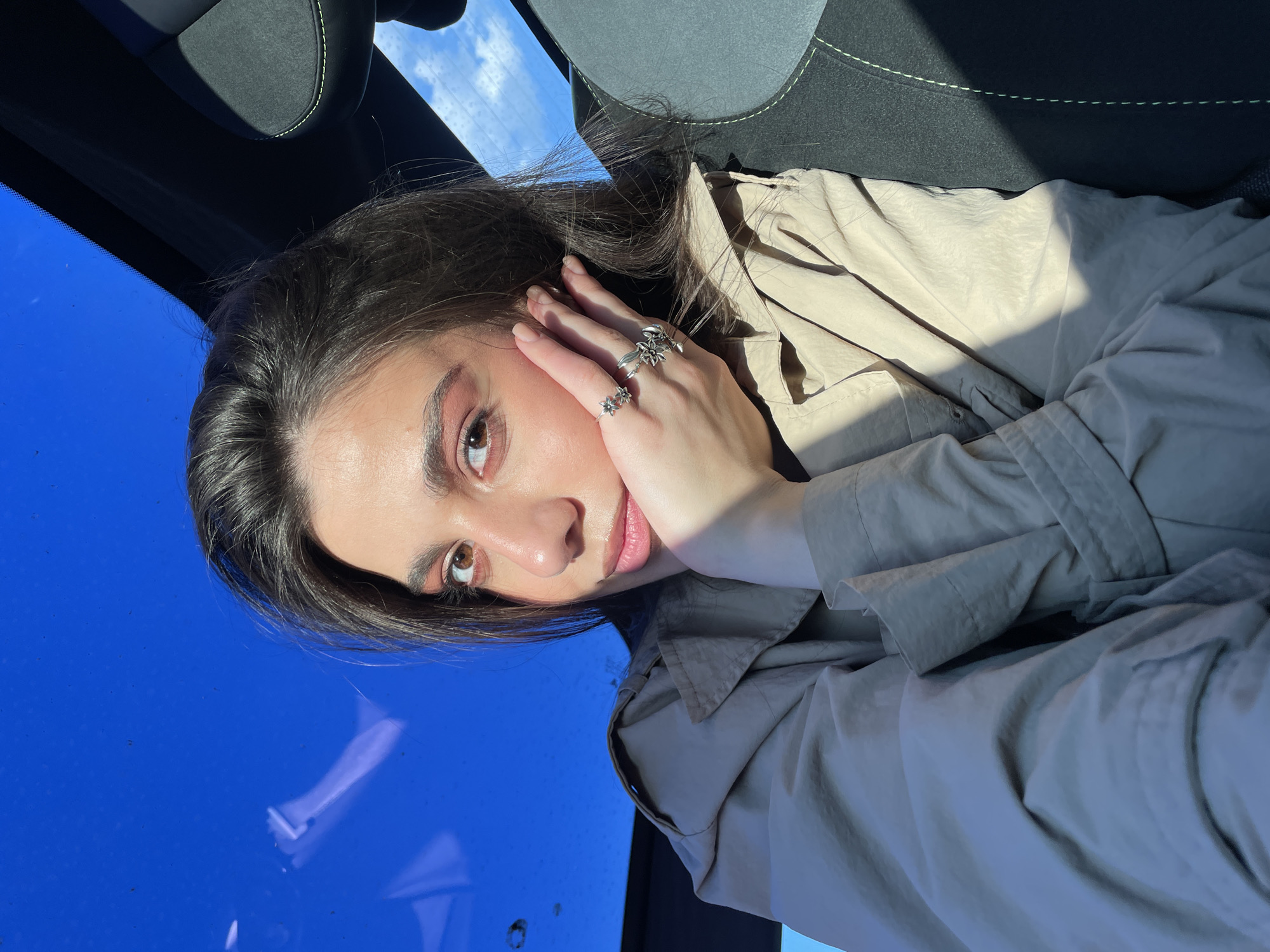 The original image captures a young woman in a close-up portrait, oriented sideways with her head on the left and her body extending towards the right. Describing it as if she were upright, she is seated inside what appears to be a car, looking directly at the viewer with a calm and composed expression. She has long, dark hair, brown eyes, and fair skin. She is wearing a light-colored, possibly beige or olive-toned, trench coat or jacket. Her left hand, adorned with multiple rings featuring intricate, nature-themed designs such as flowers, rests softly against her cheek. Behind her, a dark car seat with light-colored stitching is partially visible. Through the car's window, a bright, clear blue sky with sparse white clouds is visible, indicating a sunny daytime setting. Strong natural light casts distinct shadows across her face and the folds of her clothing. No text is visible in the image. The scene is set in Apaga, Armenia.