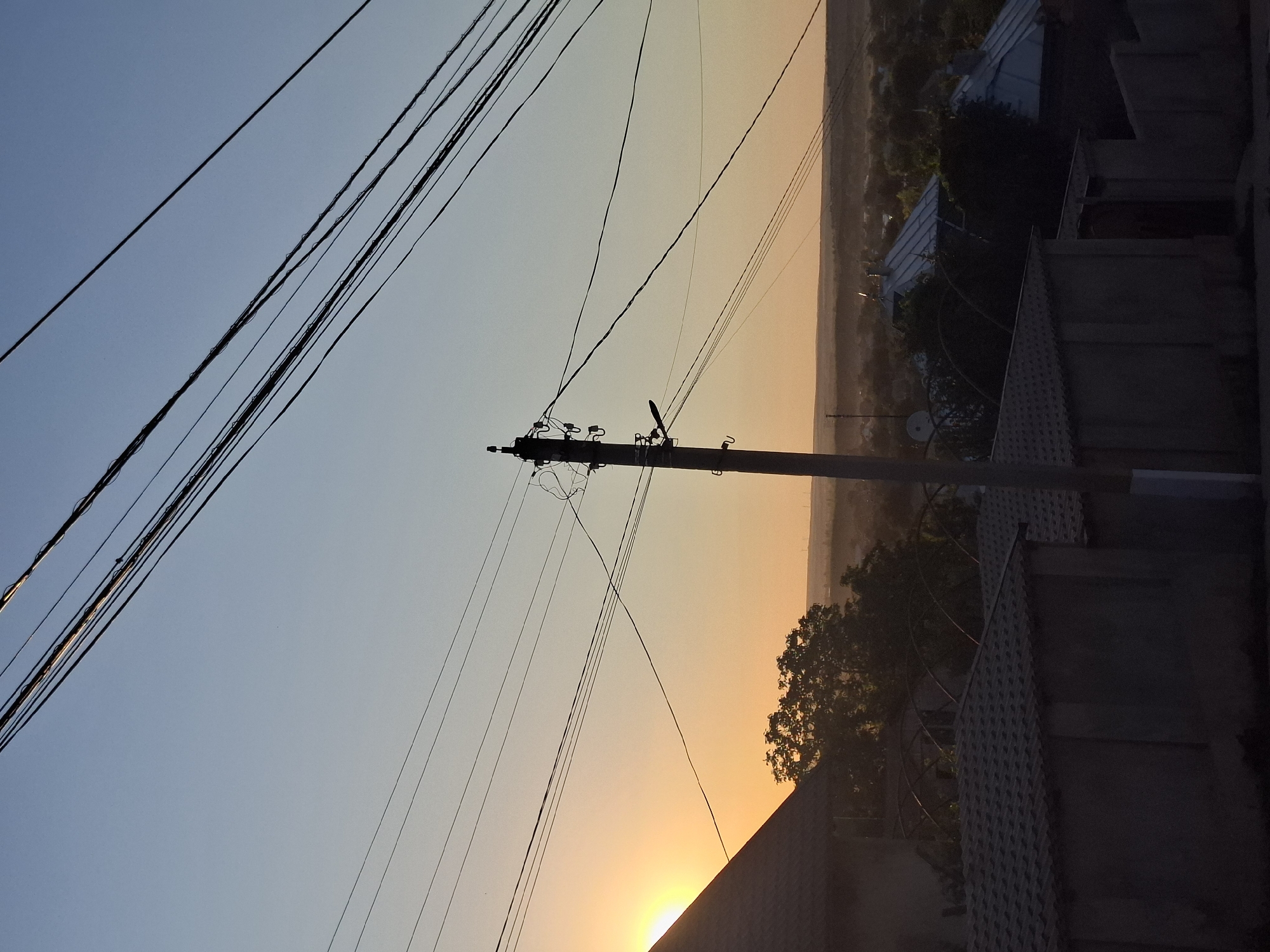 A low-angle, eye-level, medium outdoor shot captures a scene at sunset in Dubăsarii Vechi, Moldova. The sky is a gradient of light blue to pale orange, with the setting sun visible as a bright, hazy orb behind a dark, triangular roof in the left foreground. A tall, slender utility pole with numerous wires stretching across the frame is centrally located. In the foreground, a low, grey, concrete fence with a tiled roof runs horizontally. Behind the fence, several dark, peaked roofs of houses are visible, interspersed with trees. The distant landscape is a hazy expanse of rolling hills and vegetation. The overall mood is serene, with the setting sun casting a warm glow over the scene.