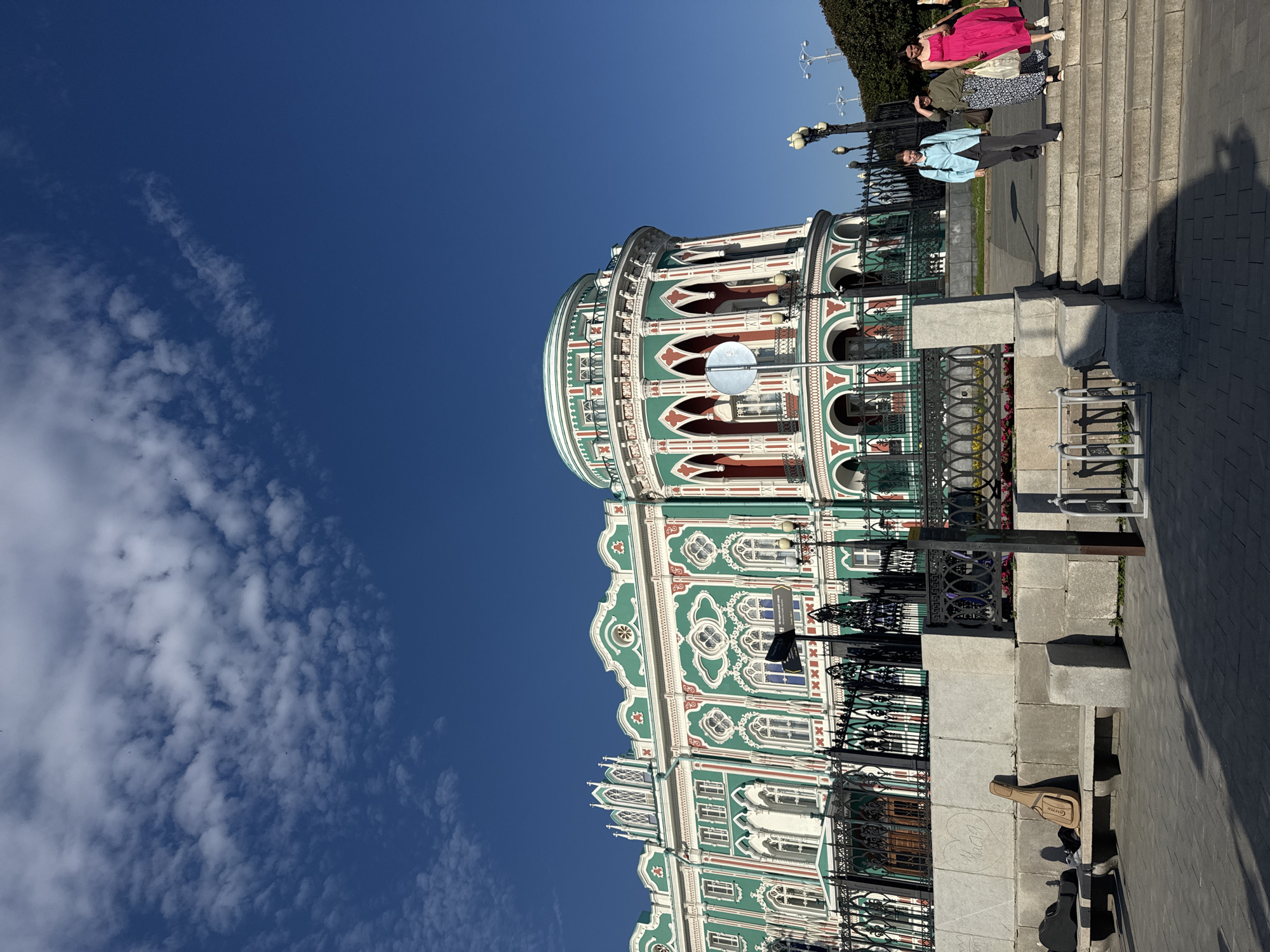 The image shows a street scene in Yekaterinburg, Russia. The foreground features a street musician with a guitar case and a guitar leaning against a stone bench. The musician is dressed in casual clothing, and the scene suggests a sunny day. Behind the musician is a decorative metal fence and a large, ornate building with green and red accents and white detailing. The building has arched windows and a domed roof, indicating it is likely a historical or cultural landmark.
In the background, several people are walking up a set of stone stairs. One person is wearing a bright pink dress, another a green jacket, and a third a blue jacket. They appear to be casually dressed and are walking away from the camera. The sky is a clear, deep blue with scattered white clouds, suggesting fair weather. The lighting indicates it is daytime, likely late morning or afternoon, with strong shadows cast by the building and people. The overall impression is of a vibrant urban environment on a pleasant day, with a blend of everyday activity and architectural beauty. No discernible text is visible in the image.