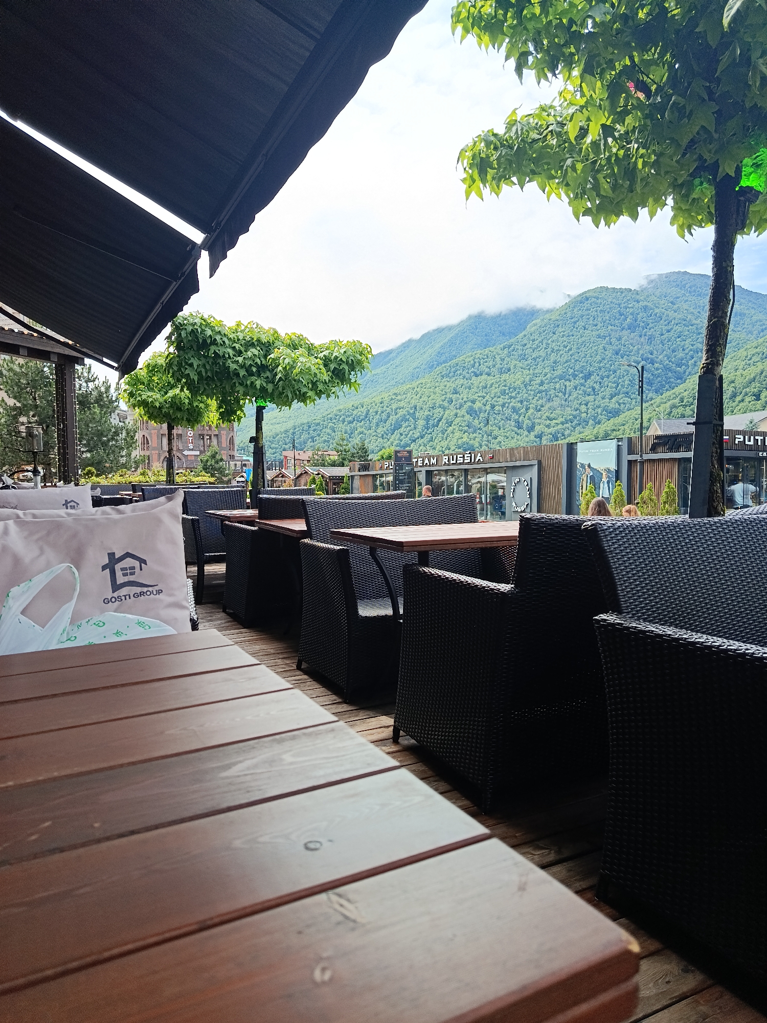 The image depicts an outdoor seating area at a cafe or restaurant with a view of forested mountains in Estosadok, Russia. The foreground is dominated by a wooden table and chairs made of dark woven material. On one of the chairs, a light-colored pillow with a house logo and the text "GOSTI GROUP" is visible, along with a white plastic bag. In the background, several tables and chairs are arranged, with patrons seated at some of them. Behind the seating area, there are buildings with signage, including "PUTIN TEAM RUSSIA". The mountains in the distance are covered in lush green trees, with some clouds obscuring the peaks. A large tree with green foliage is prominently featured in the middle ground, and another tree with a glowing green light is visible to its right. The overall scene suggests a relaxed atmosphere in a scenic mountain resort town.