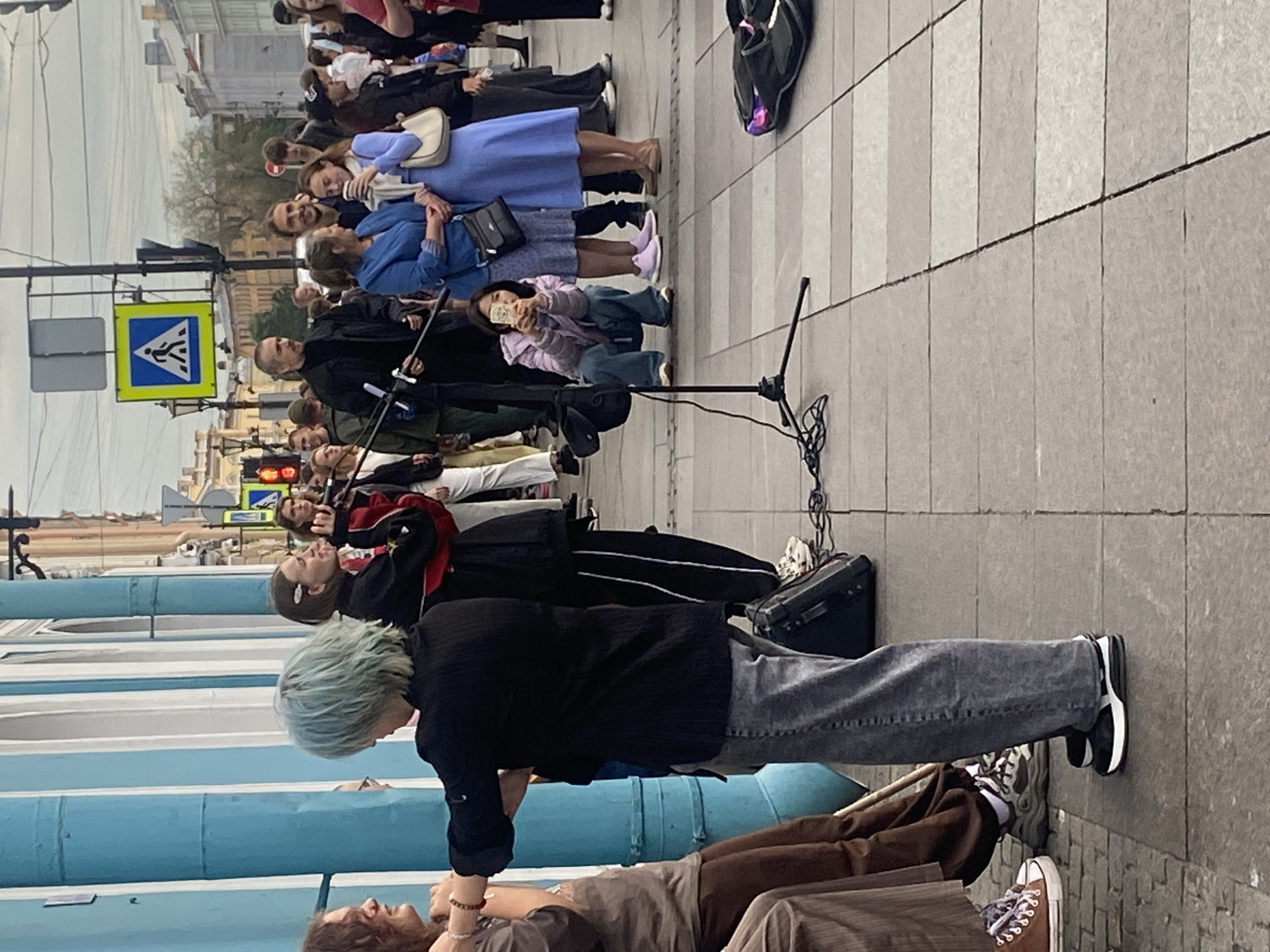 The image depicts an outdoor scene in Saint Petersburg, Russia, likely on a pedestrian street. In the foreground, several individuals are gathered. A person with light blue hair is standing, facing away from the camera, holding a microphone stand. To their left, another person is partially visible, wearing a brown outfit and brown sneakers. Another person in the foreground is wearing grey jeans and black and white sneakers. A microphone and speaker are set up on the pavement, suggesting a performance or street event.
In the background, a group of people are visible, some interacting with each other. There is a traffic light and a pedestrian crossing sign overhead, indicating an intersection nearby. Buildings with classical architecture, typical of Saint Petersburg, line the street. The sky is overcast. The overall impression is a lively street scene with a focus on a performance or gathering.
