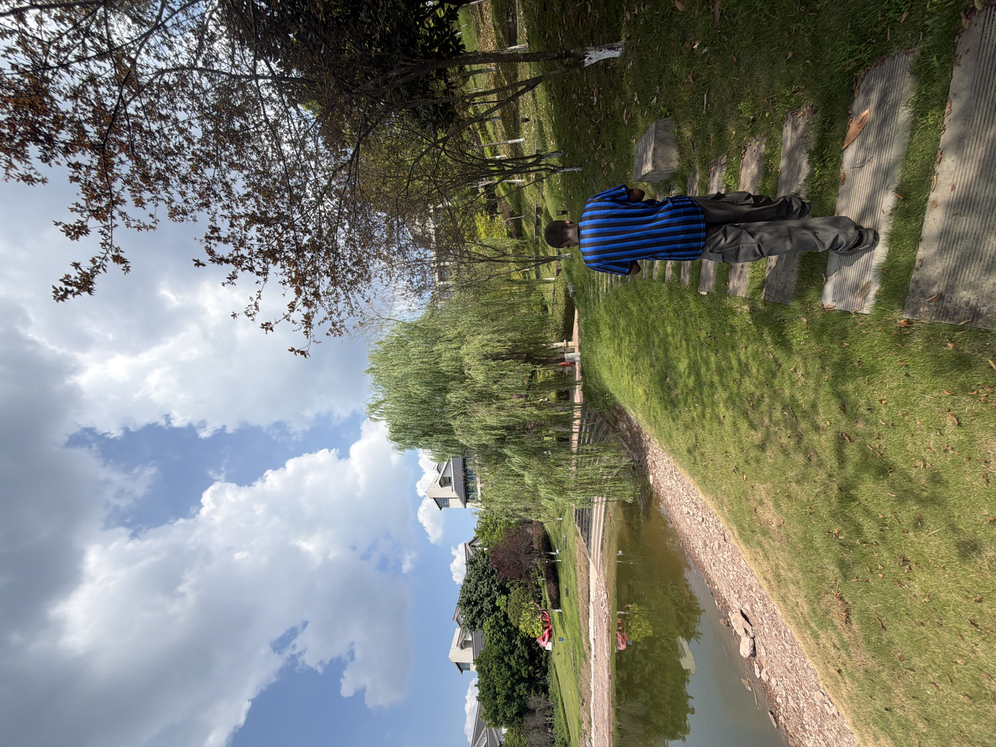 A person, from behind, walks on a stone path in a park with a lake and buildings. The person is wearing a blue and black striped t-shirt and gray cargo pants. The path is surrounded by green grass and trees. In the background, there is a lake with reflections of trees and buildings, and several modern buildings are visible on a hill. The sky is partly cloudy with blue sky peeking through. The image appears to be taken in the city of Jiangjin, China, possibly at a park or residential area with landscaped grounds.