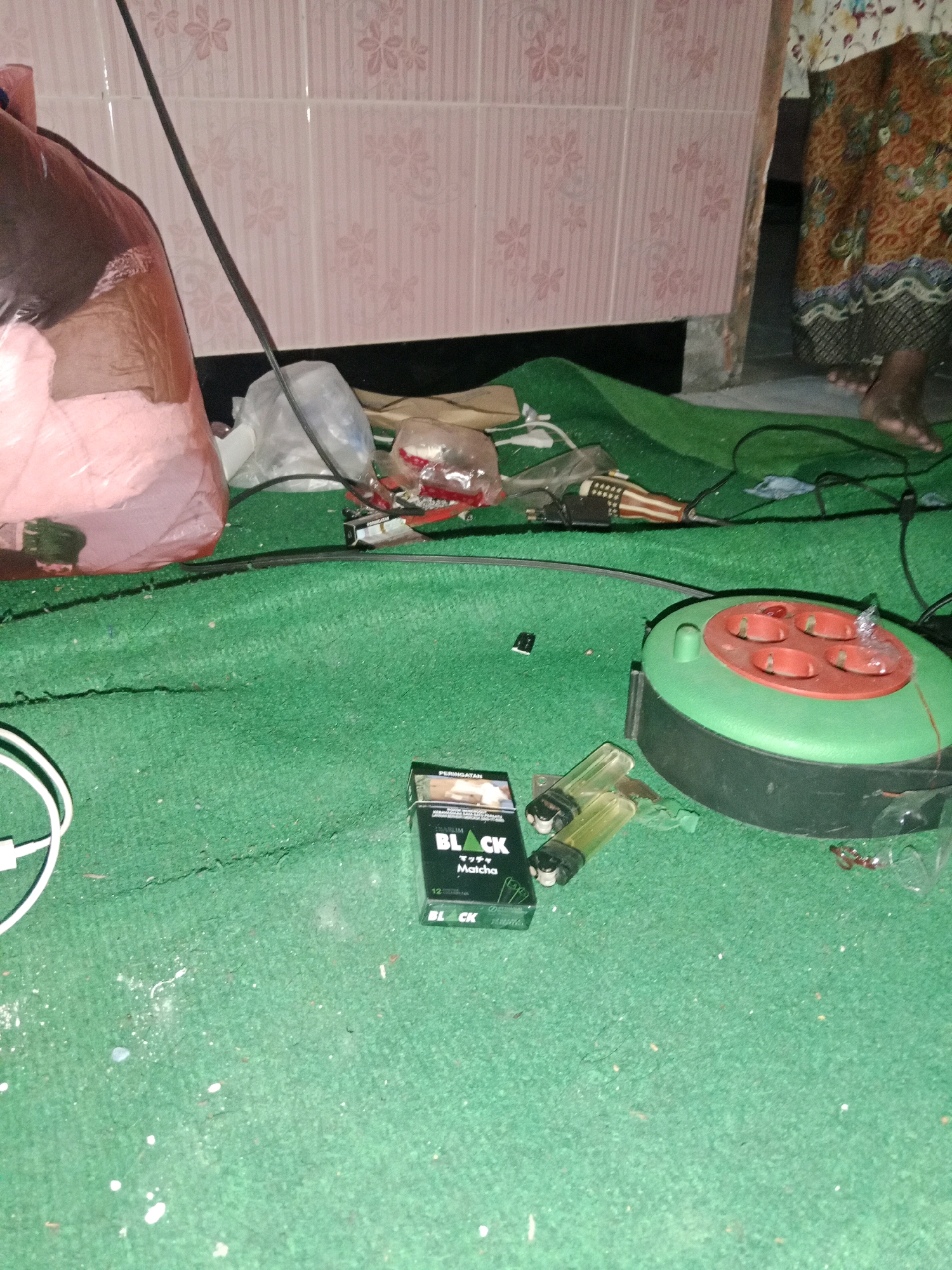 The image depicts a close-up view of a green carpeted floor, likely indoors. Several objects are scattered across the carpet, suggesting a cluttered environment. In the foreground, a pack of "BLACK" cigarettes and two lighters are visible. To the right, a green and red electrical extension cord reel is present. In the background, a large pink plastic bag, possibly containing laundry or other items, is partially visible. A black wire runs diagonally across the scene. The wall behind the objects appears to be tiled with a pink floral pattern. The lower right portion of the image shows a person's legs and feet, wearing a patterned skirt and appearing to be standing or walking. Based on the context provided, this scene is located in the city of Ringinrejo, Indonesia.