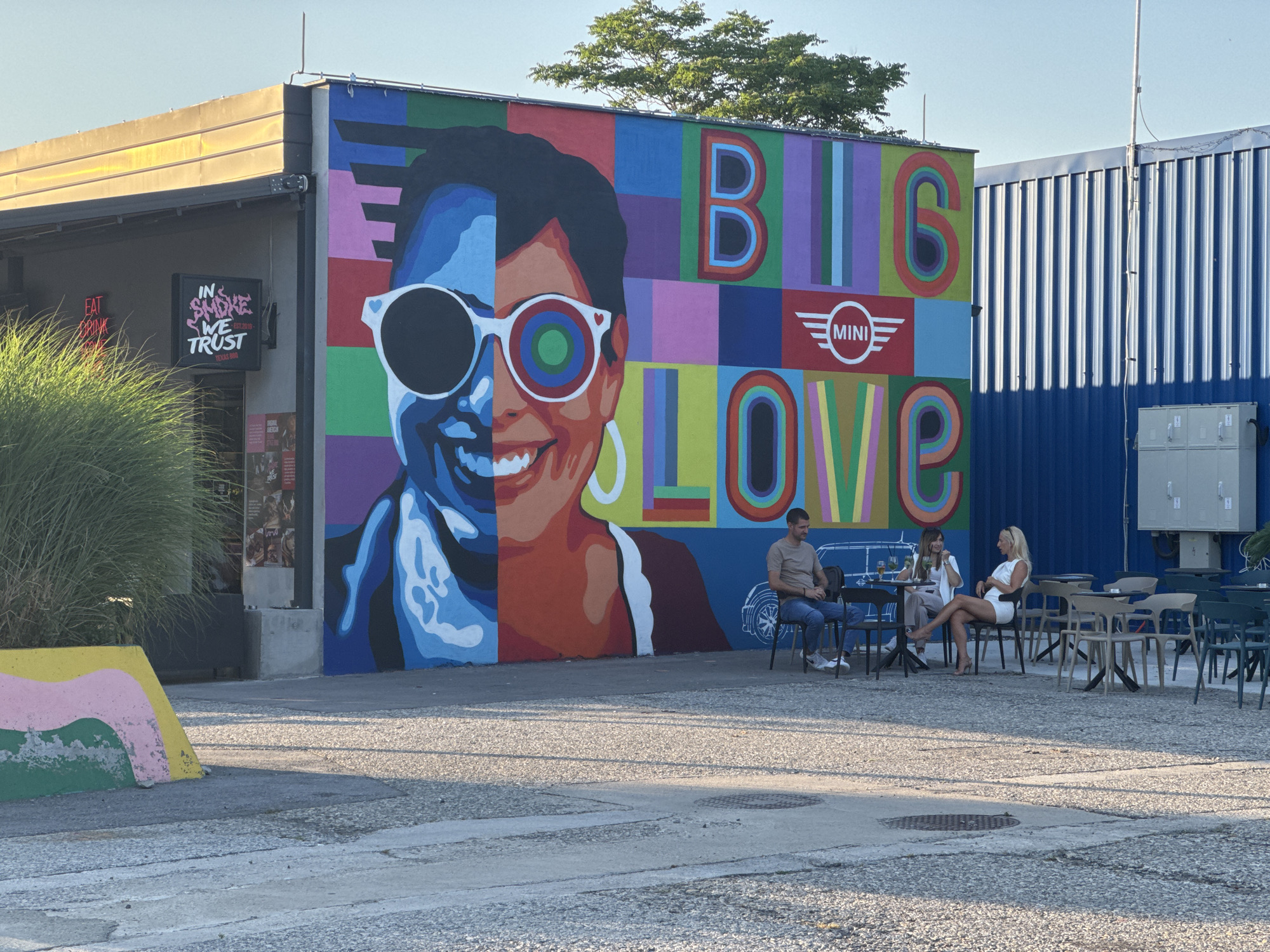 This media file captures an outdoor scene with a vibrant mural as the backdrop. The mural depicts a large portrait of a person, split down the middle with contrasting colors and styles, and features the words "BIG LOVE" and a MINI logo. In front of the mural, several people are seated at tables, appearing to be at an outdoor cafe or restaurant. A man in a light brown t-shirt and jeans sits at a table with two women. One woman is wearing a white top and light-colored pants, while the other is in a white dress and high heels. They are all seated and appear to be engaged in conversation, with drinks on their tables.
To the left of the mural, part of a building is visible with signage that includes neon pink text "EAT DRINK" and a black sign with "IN SMOKE WE TRUST EST. 2019" in pink. A large clump of green grass or foliage is situated in front of this building. The ground is paved with concrete and gravel. In the background, there are more tables and chairs, as well as a tall blue corrugated metal structure. The lighting suggests it is daytime, likely late afternoon or early evening, given the warm sunlight casting shadows. The overall atmosphere appears relaxed and social. The location is identified as Belgrade, Serbia.