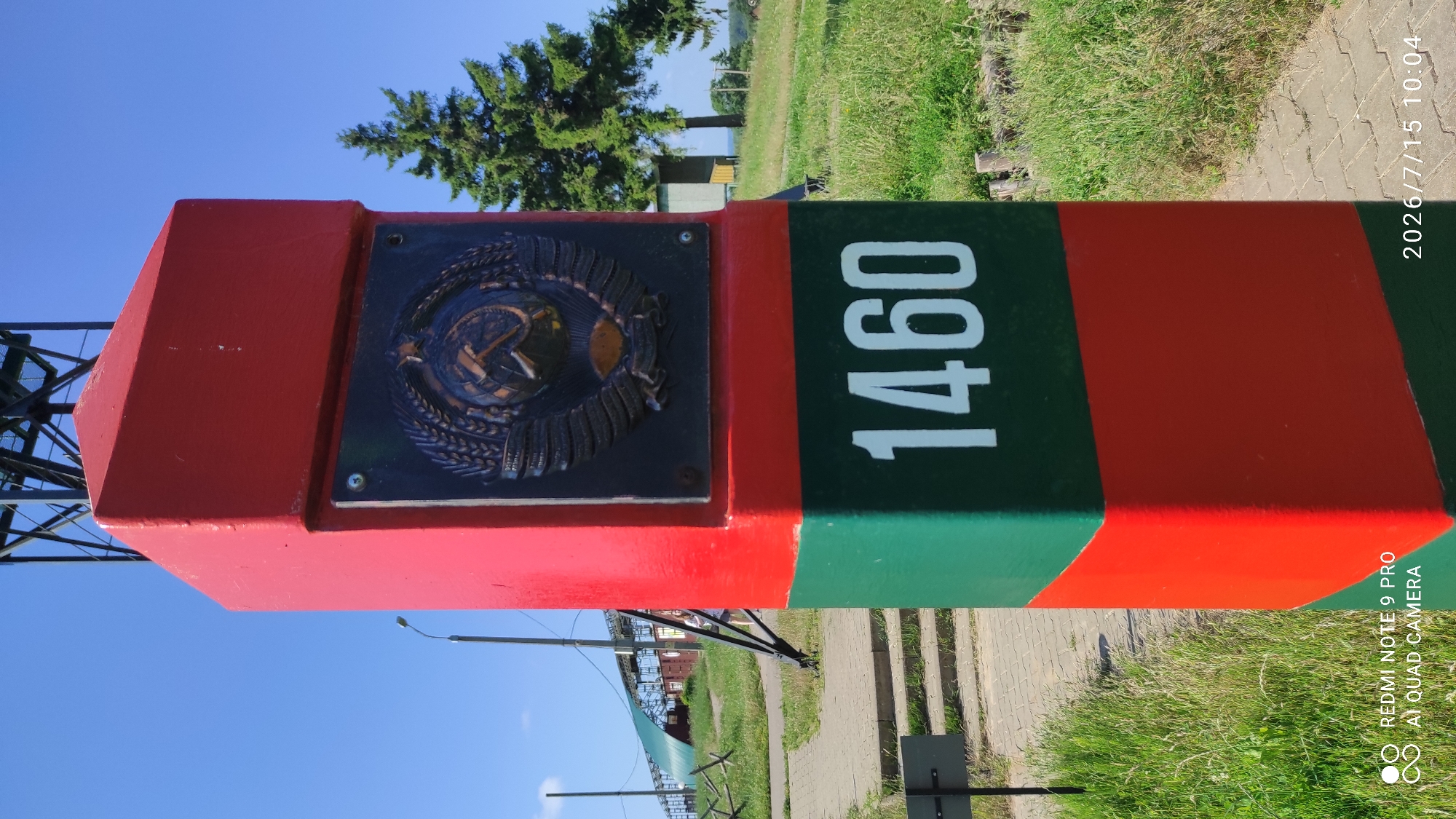 A close-up, eye-level outdoor shot features a red and green striped concrete border post in the foreground, set against a bright blue sky. The post is topped with a red cap and displays a circular Soviet emblem, featuring a hammer and sickle within a wreath of wheat, affixed to its upper section. The number "1460" is painted in white on the green band of the post.
The post stands on a paved pathway made of interlocking concrete blocks, with a grassy area to its right and left. In the background, to the left, a woman wearing a light-colored dress is partially visible walking away from the camera. Behind her are some industrial-looking structures, including what appears to be a green awning or covering and some metal supports. To the right of the post, a tall, dark green coniferous tree is prominent, with a small, light-colored building visible behind it. Further in the distance, the landscape consists of rolling green hills under a clear blue sky. The overall scene appears to be a border crossing or a historical marker site. The timestamp in the lower right corner indicates July 15, 2026, at 10:04 AM. The text "REDMI NOTE 9 PRO AI QUAD CAMERA" is present in the lower left corner.