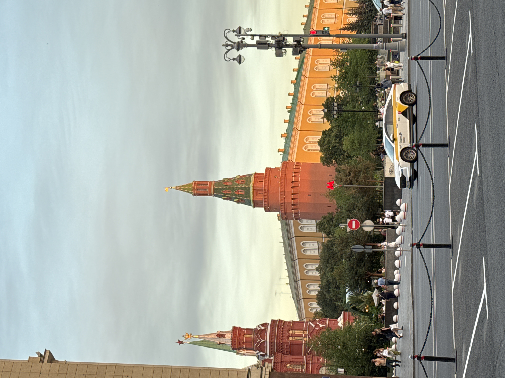 This image depicts a street scene in Moscow, Russia, likely near Red Square, given the architectural style of the towers. In the foreground, a street is visible with white lane markings and black stanchions connected by chains, delineating pedestrian areas from the road. A white taxi with yellow accents and Russian text, possibly identifying it as a Yandex Taxi, drives on the road.
In the middle ground, several people are walking and standing on a sidewalk. A red and white traffic sign with a red circle and horizontal bar, indicating a prohibition, is also present. Behind the sidewalk is a row of decorative white planters. Lush green trees are scattered throughout this area, providing shade and separating the street from the buildings.
The background is dominated by a large, ornate building complex, characterized by its yellow facade, numerous arched windows, and green roofs. Two prominent towers are visible to the left and center. The left tower is red with a green spire topped by a golden eagle and a red star. The central tower is taller, also red with a green, tiered spire. The lighting suggests it is either early morning or late afternoon, with a warm glow illuminating the facade of the building and the towers. The sky is overcast with a pale blue hue.